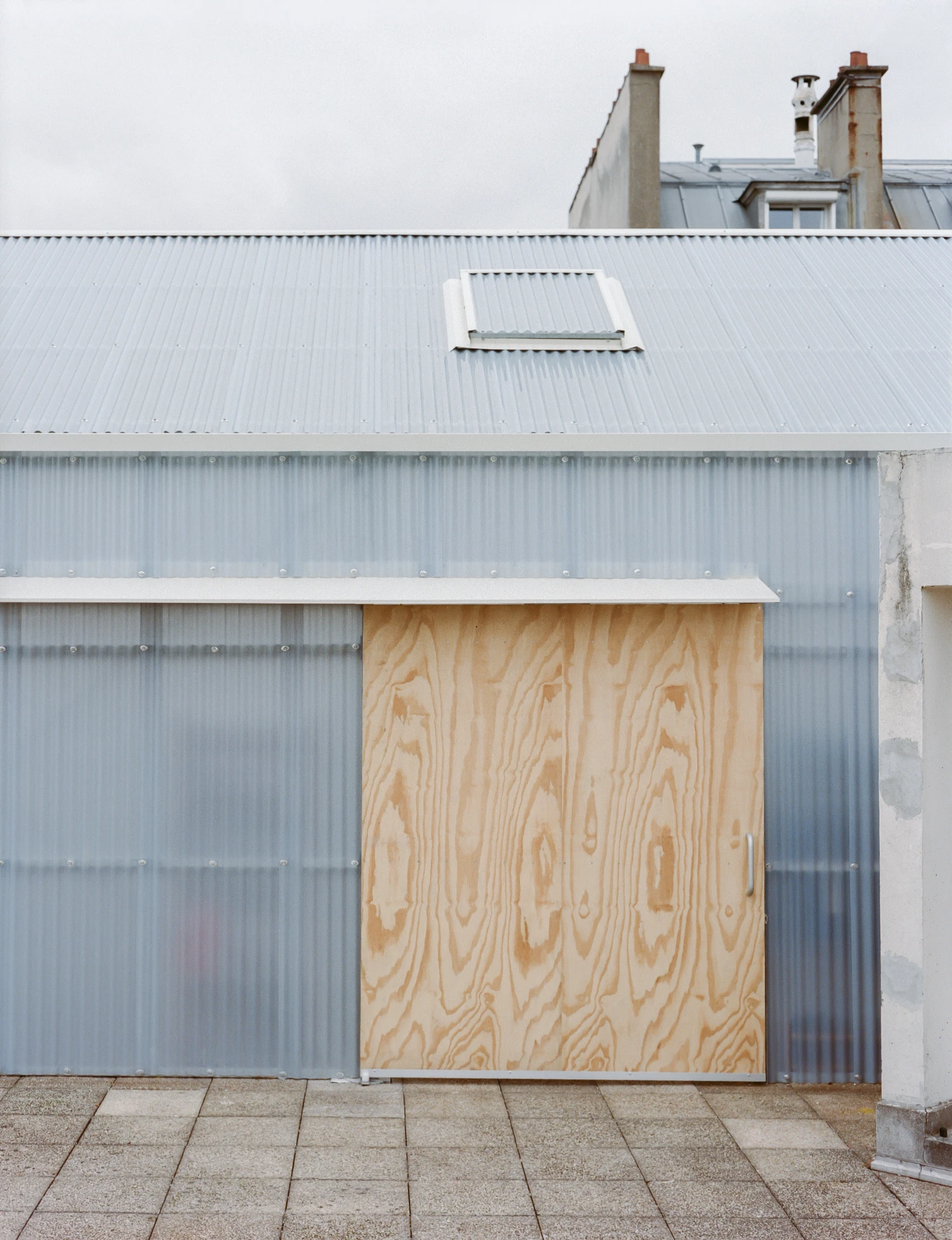 Rooftop Educational Farm by rerum architectes — A corrugated metal structure with plywood sheathing dominates the foreground, its pale timber facing punctuated by wood grain patterns.