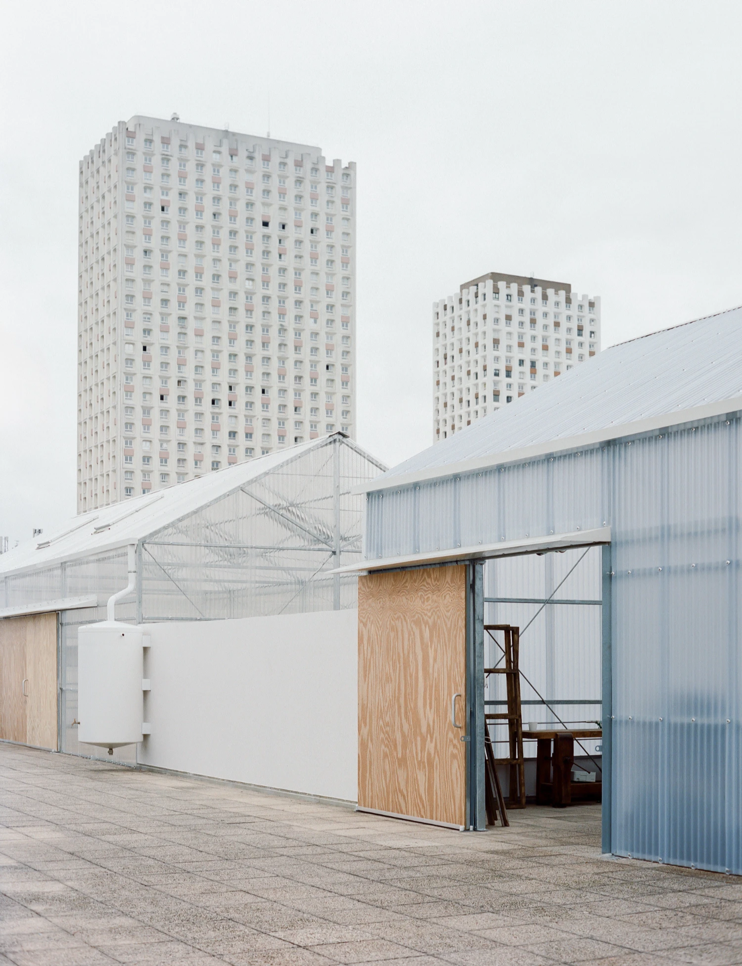 Rooftop Educational Farm by rerum architectes — A rooftop greenhouse structure on a Parisian tower block features a triangulated steel frame with diagonal bracing supporting polycarbonate panels that...