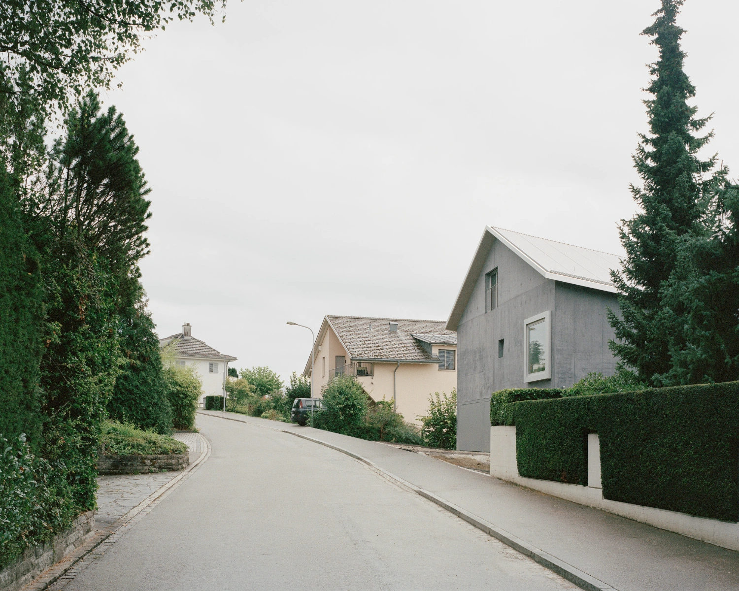 House on a Slope by KARAMUK KUO — A residential street in Zollikon frames the gray concrete facade of KARAMUK KUO's five-unit dwelling, its pitched gable roof rising prominently against a...