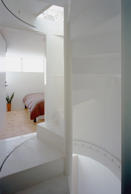 Long Tall House by Spacespace — View through white-rendered partition wall into multi-level interior, revealing stacked bedroom spaces with wooden flooring and burgundy bedding.