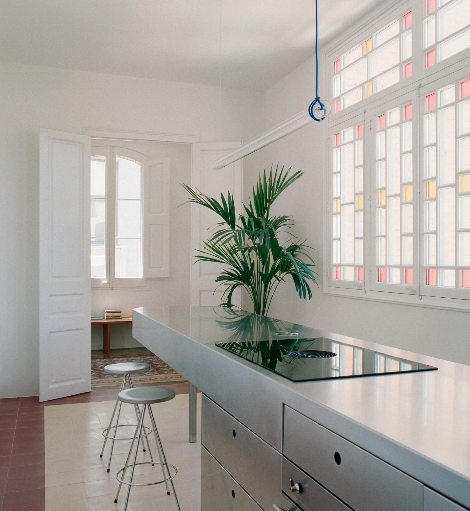 LL731 by Aramé Studio — A linear kitchen workspace anchored by a continuous white laminate counter, punctuated by black circular hardware and supporting a lush potted palm.