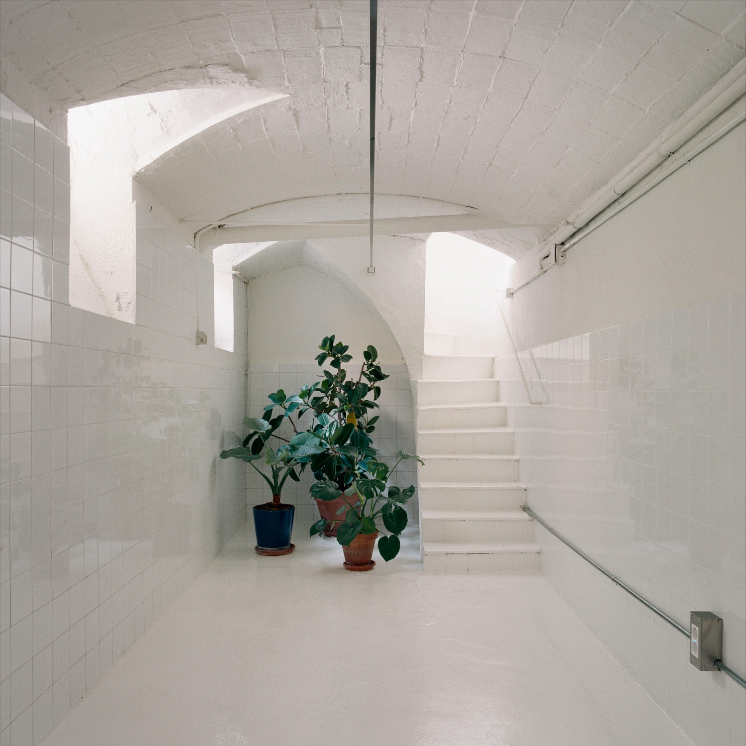 LL731 by Aramé Studio — A white-tiled corridor ascends via a cantilevered staircase with steel handrail, its risers rendered in plaster.