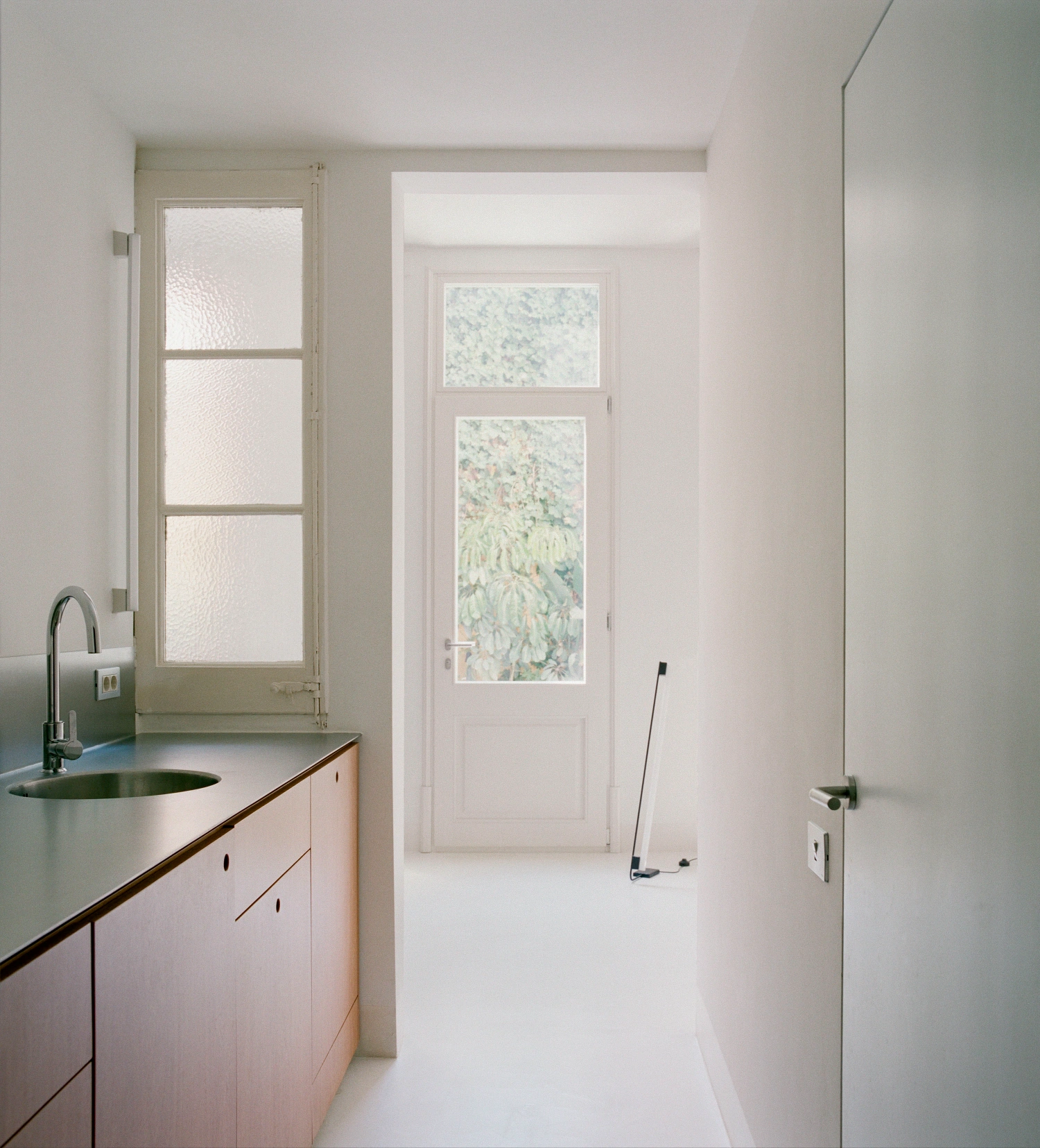 LL731 by Aramé Studio — A galley kitchen with pink-painted cabinetry and stainless steel countertop features a rounded sink positioned beneath a gridded window with translucent panes.