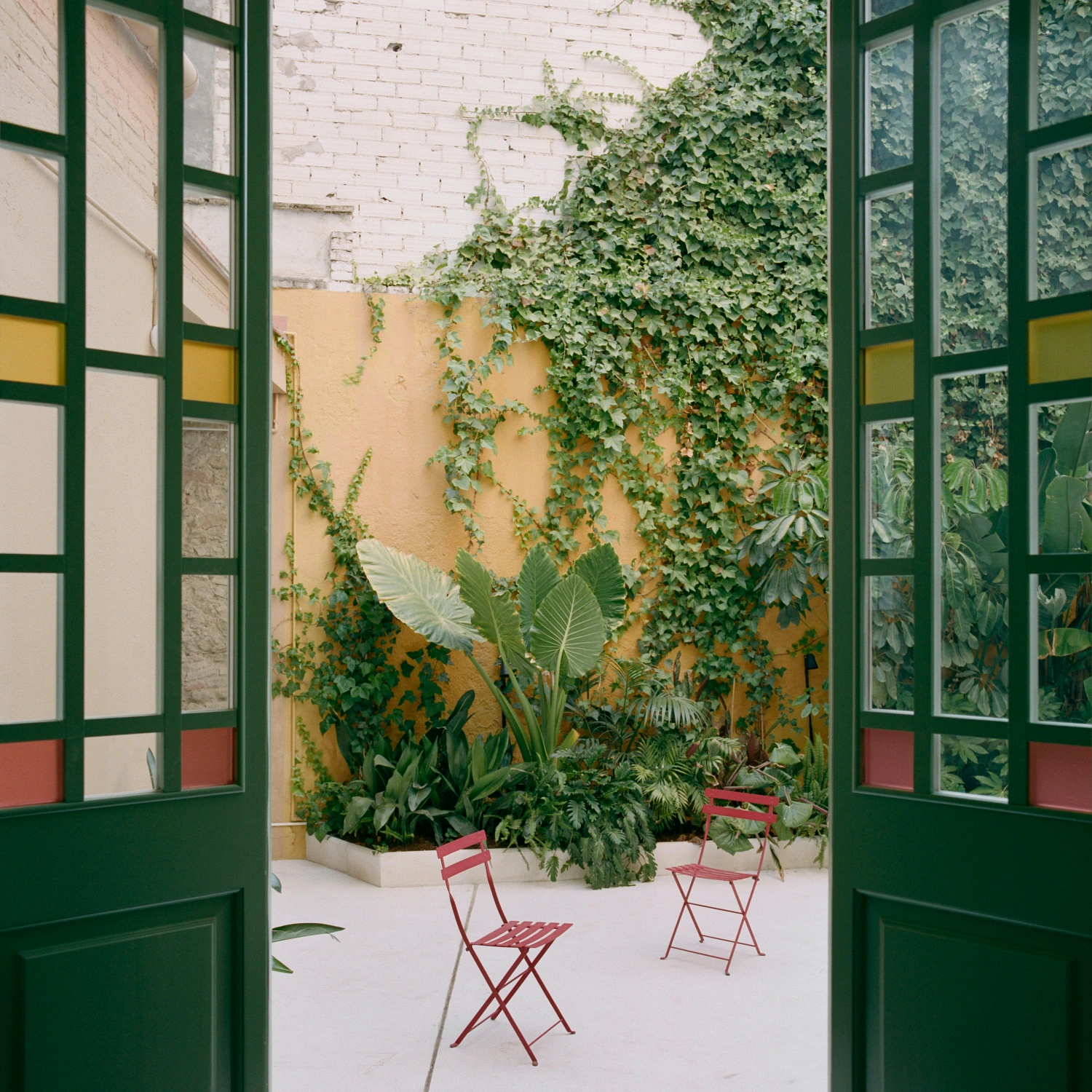 LL731 by Aramé Studio — A courtyard viewed through paired green-painted steel-frame doors with gridded glass panes and colored panels in yellow, cream, and terracotta.