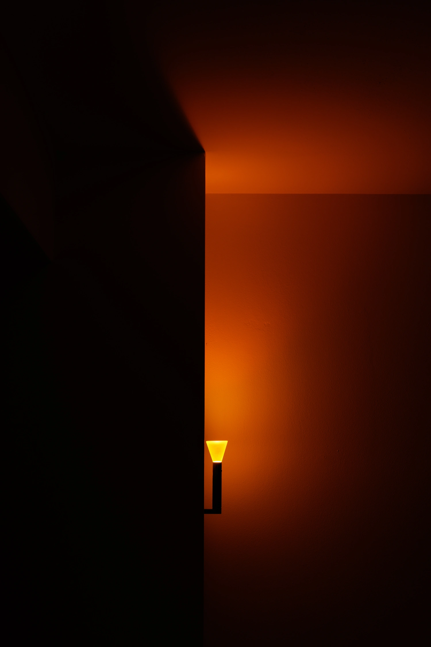 Halo Mag (Flame + Bloom) designed by Mandalaki — A minimal lamp positioned in a darkened architectural threshold, its translucent satin-finish diffuser emitting warm yellow light from a matte black...
