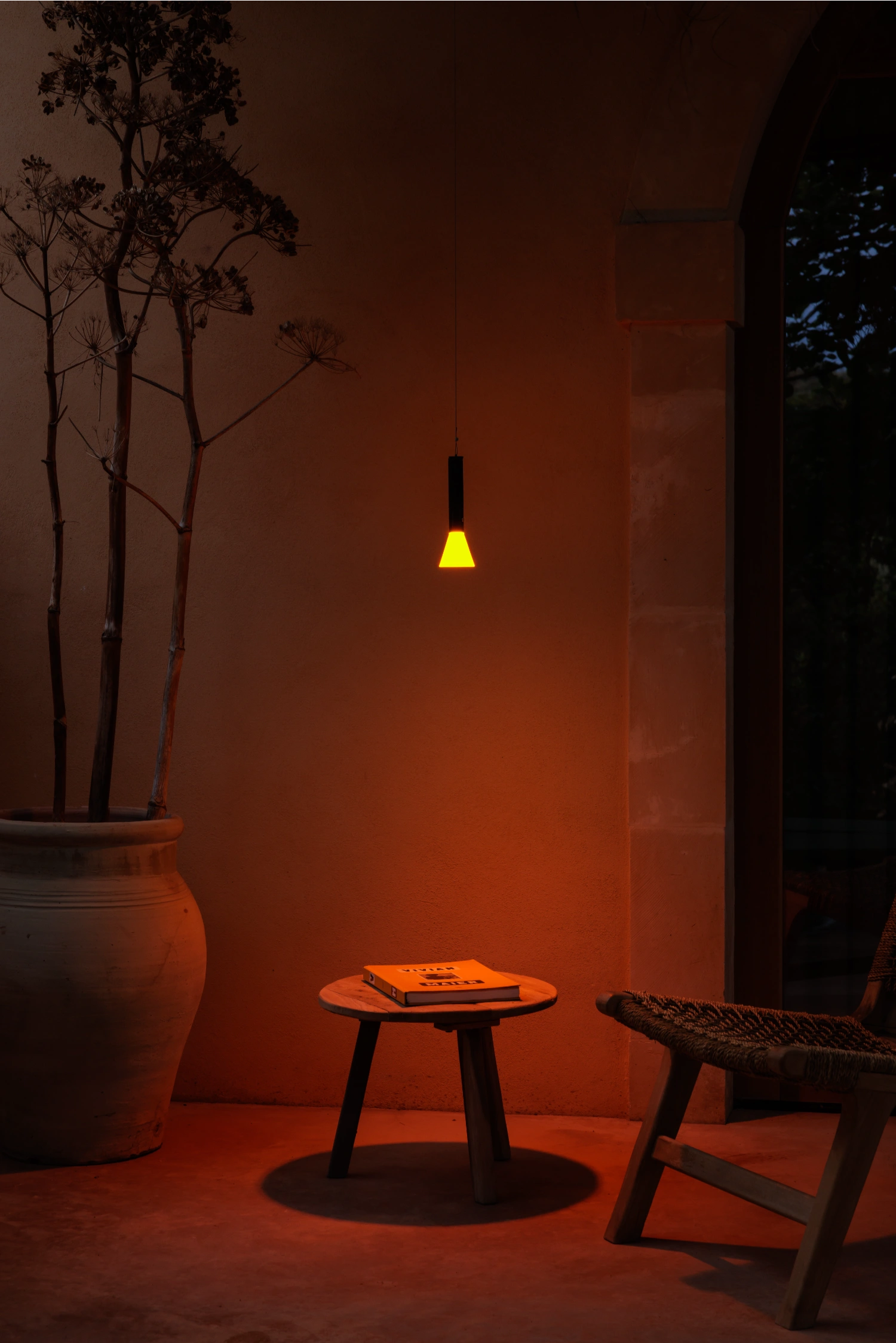 Halo Mag (Flame + Bloom) designed by Mandalaki — A pendant lamp with a translucent satin-finish diffuser in warm yellow tone hangs above a minimal black three-legged table positioned on terracotta-rendered...
