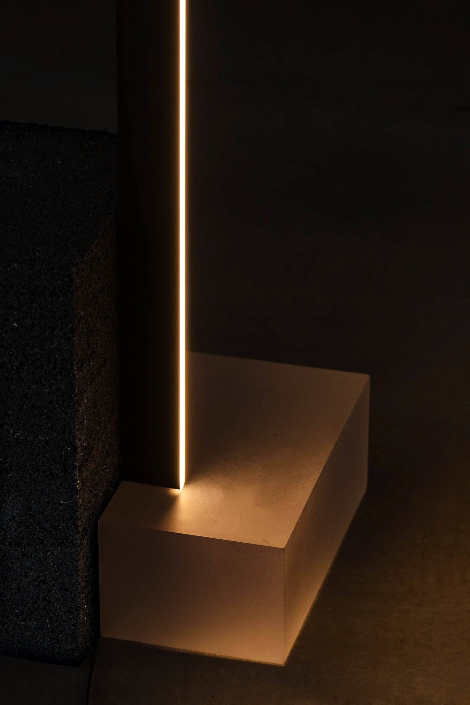 As by So Koizumi — A vertical brass or bronze rod extends upward from a cream-colored concrete block, its slender form backlit to create a luminous linear accent against darkness.