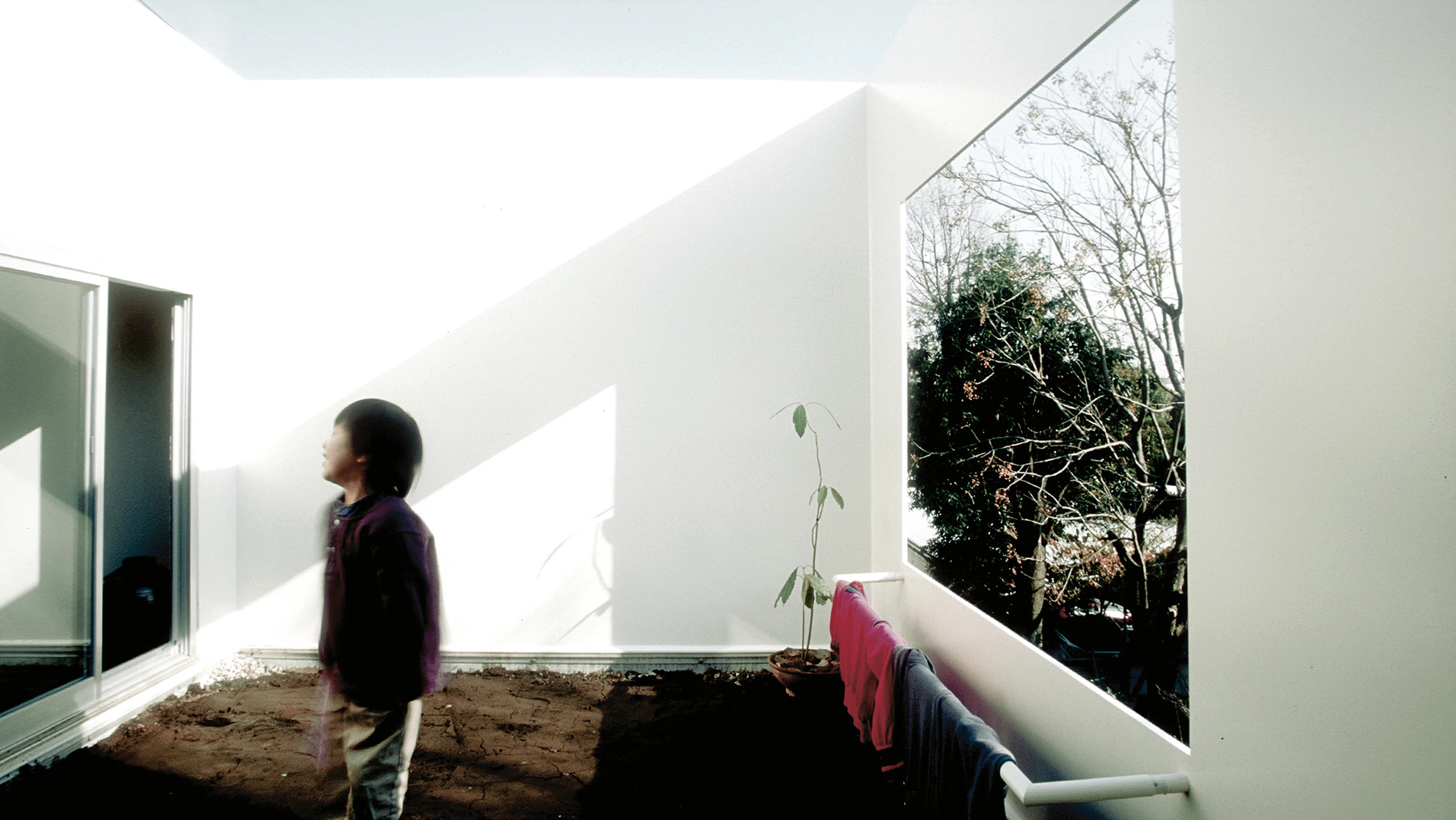 House in a Plum Grove by Kazuyo Sejima by Kazuyo Sejima & Associates — A child in burgundy outerwear stands within the compressed exterior courtyard of Sejima's plum grove house, oriented toward the recessed entrance.