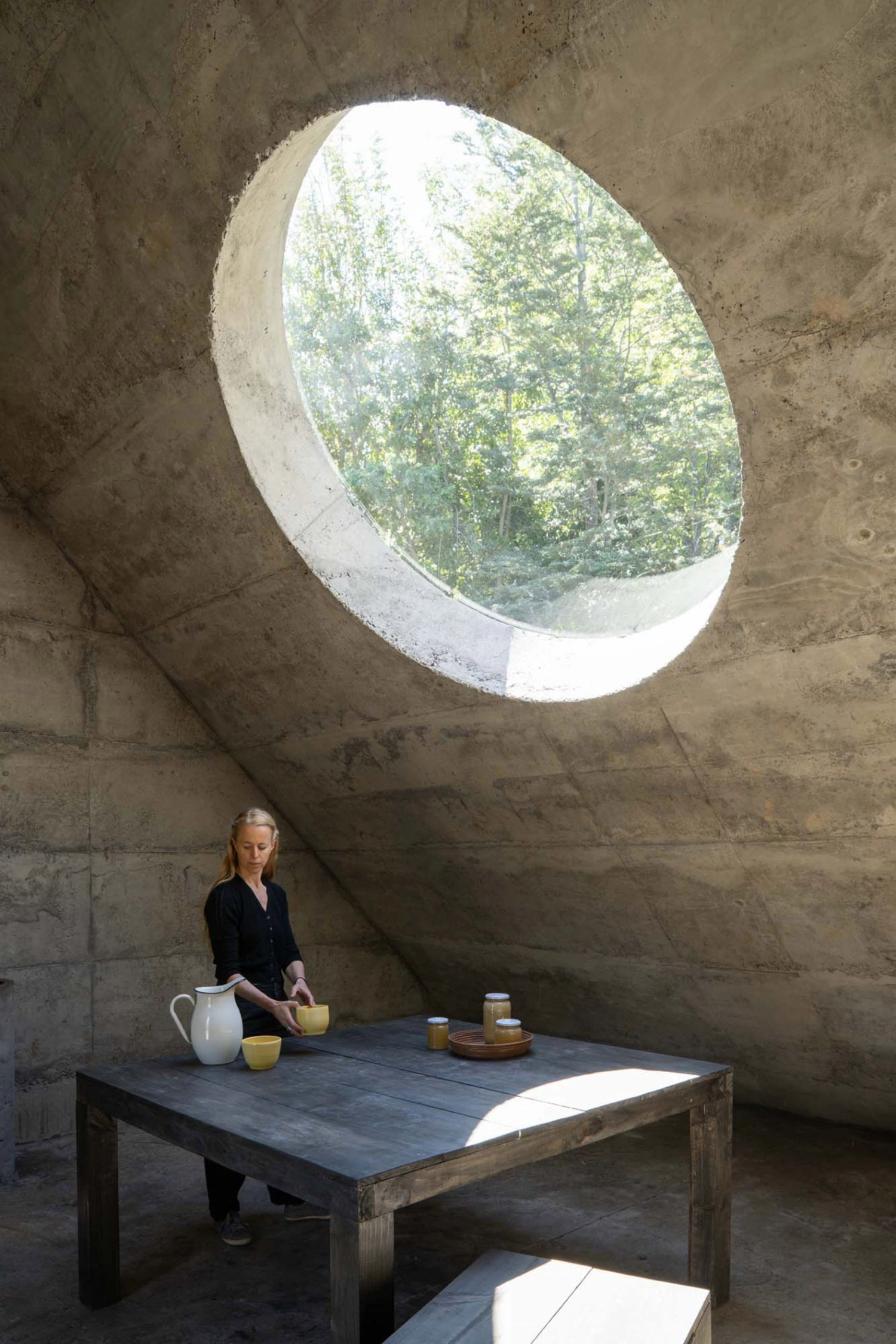 MIEL Pavilion by Pezo von Ellrichshausen — A woman stands at a timber table within the concrete volume of MIEL Pavilion, positioned beneath a large circular aperture that frames dense forest canopy...
