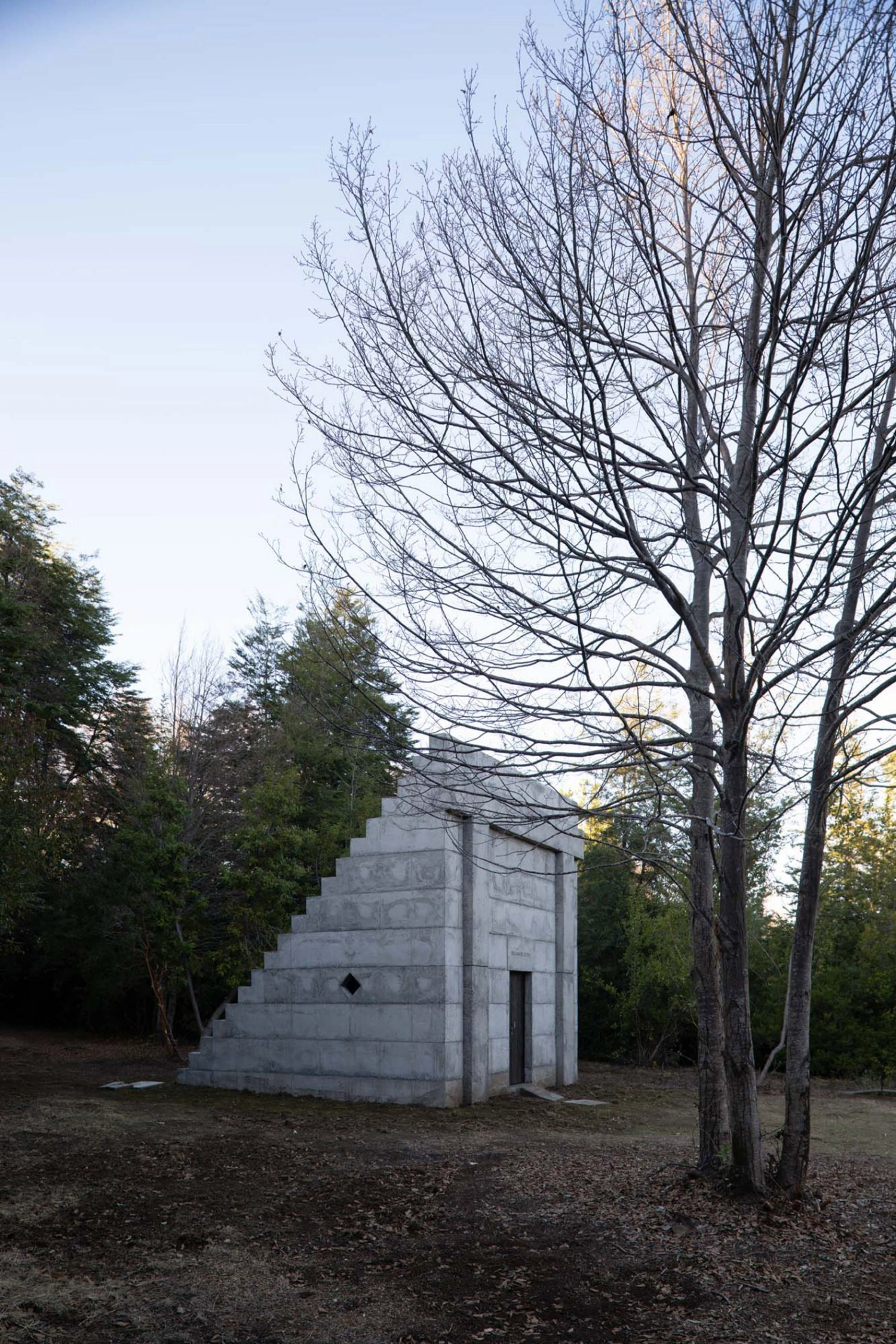 MIEL Pavilion by Pezo von Ellrichshausen — The MIEL Pavilion appears as a monolithic concrete structure set within a woodland clearing, its stepped facade rising against bare deciduous trees.
