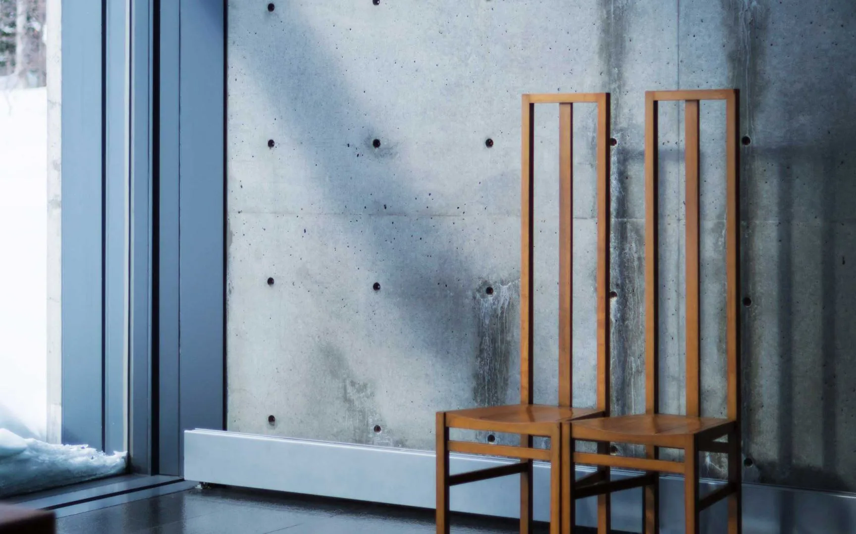 The Church on the Water by Tadao Ando — Two tall wooden chairs with vertical slat backs occupy the foreground of this interior view, positioned symmetrically before a raw concrete wall.