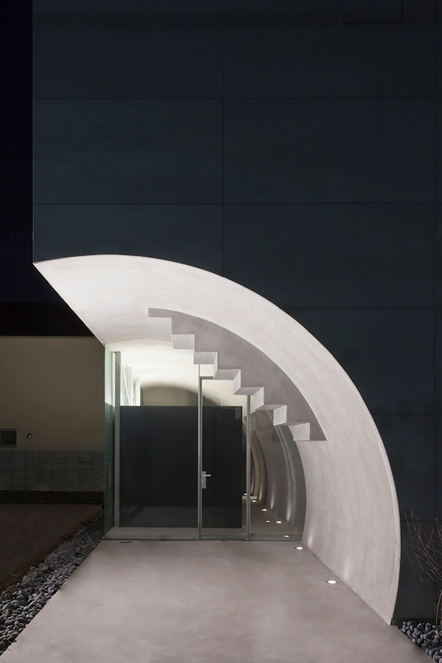Tunnel House by Makiko Tsukada Architects — The Tunnel House entrance presents a quarter-cylinder void carved through the building's facade, its curved white concrete walls creating a luminous passage...
