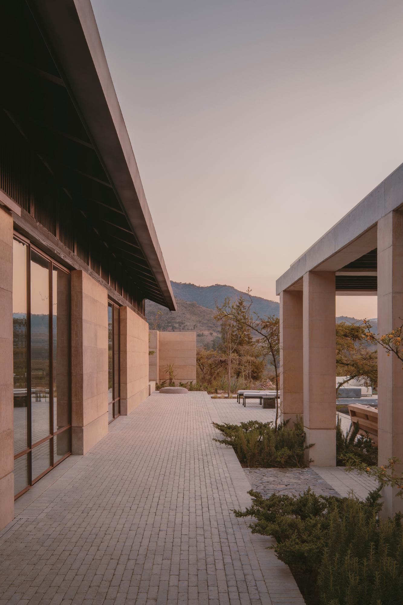 Casa el Espino by Soler Orozco Arquitectos — A brick-paved courtyard extends between terracotta-rendered walls and white concrete columns, framed by the receding perspective of the covered passage.