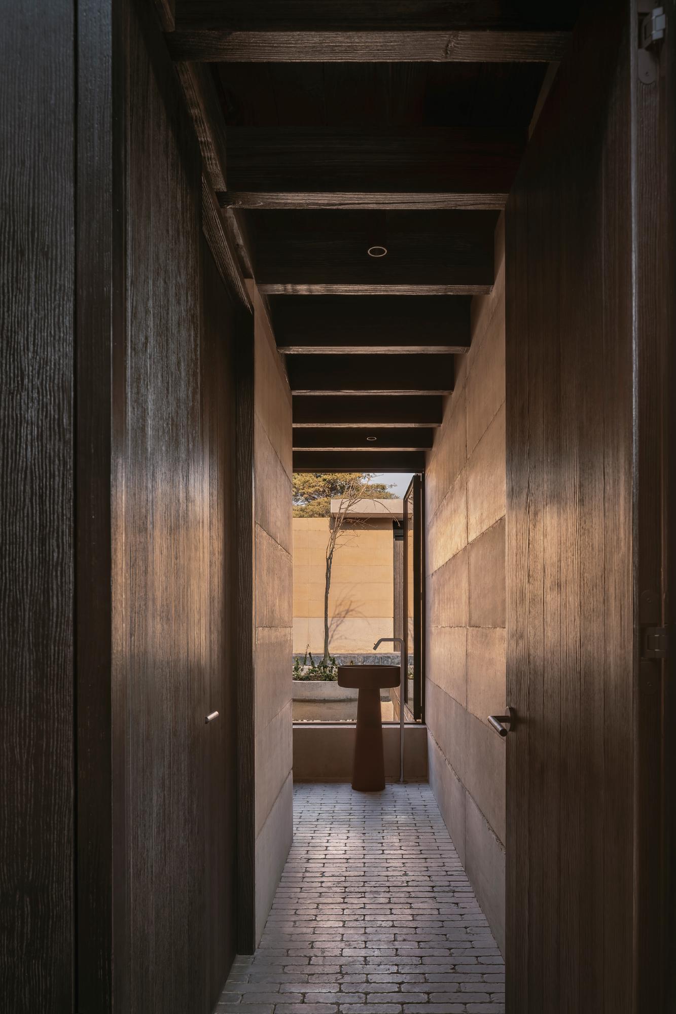 Casa el Espino by Soler Orozco Arquitectos — A timber-lined corridor frames the passage toward a sunlit courtyard, its perspective compressed by dark wood-clad walls on either side.