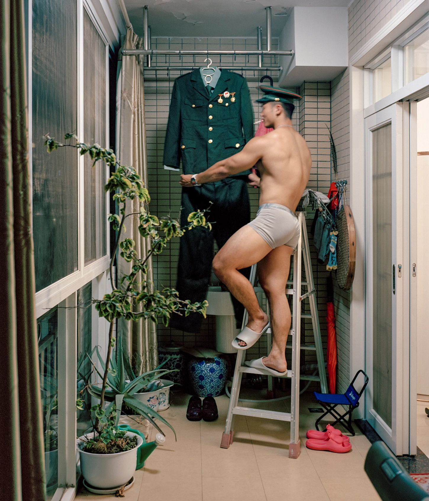 The Internal Crusade by Zexuan Zeng — A shirtless man stands on a metal ladder in a narrow residential corridor, reaching toward a dark green military jacket displayed on a wall-mounted garment...