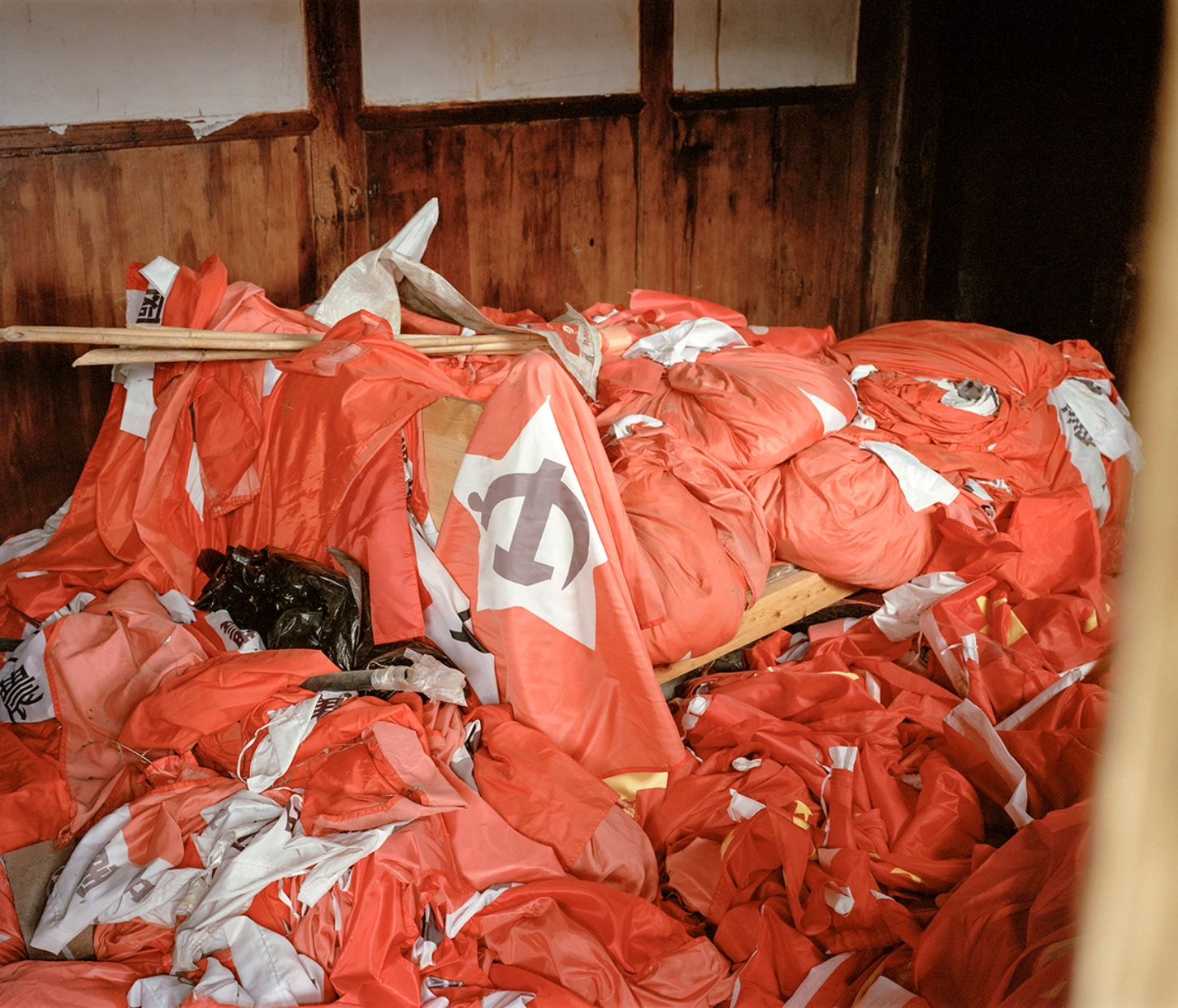 The Internal Crusade by Zexuan Zeng — A warehouse interior accumulated with discarded red and white nylon jackets bearing graphic prints, piled across wooden shelves and floor.