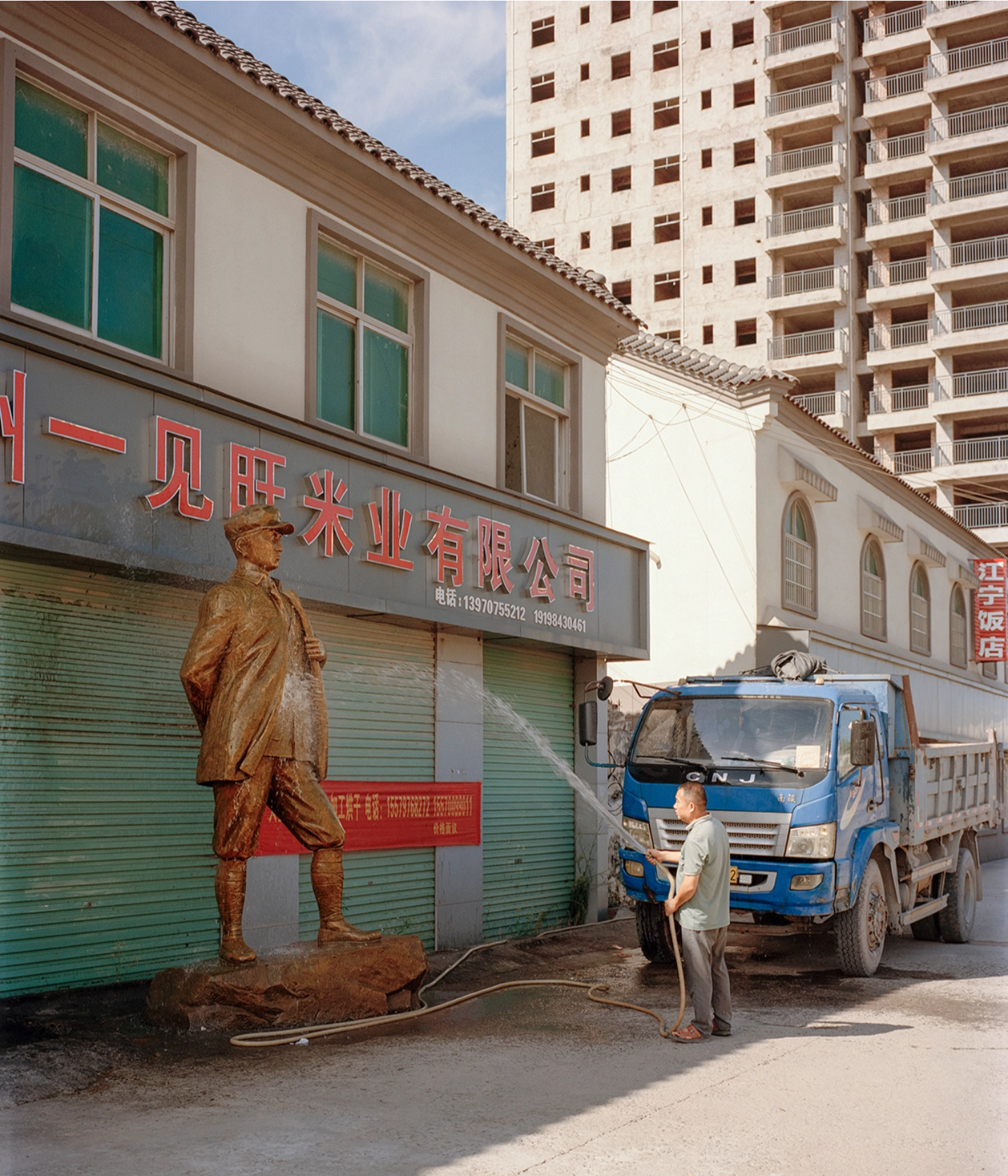 The Internal Crusade by Zexuan Zeng — A bronze statue of a revolutionary figure in military coat stands sentinel before a commercial storefront with green metal roller shutters, its surface...