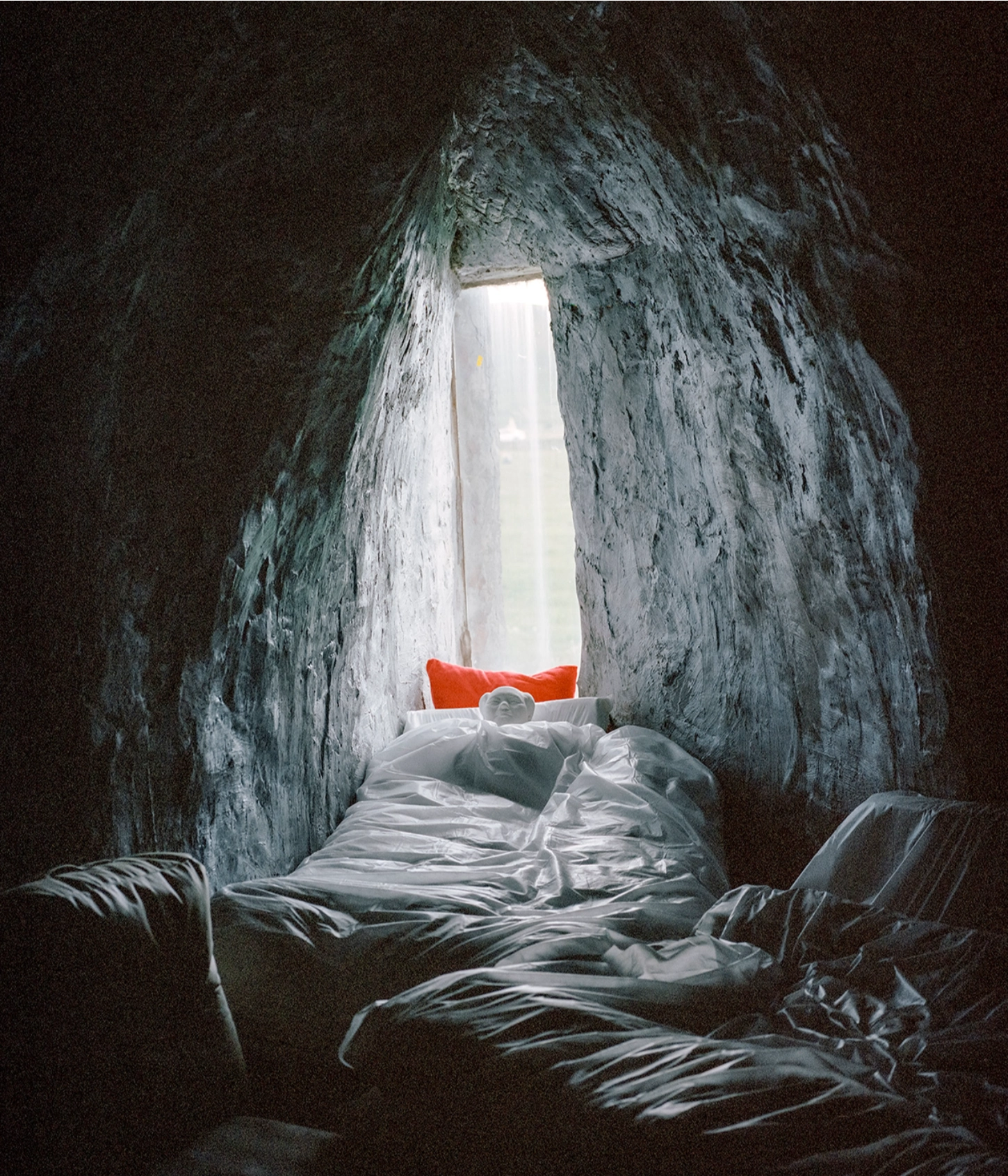 The Internal Crusade by Zexuan Zeng — A bed occupies a narrow stone tunnel, its white linens and red pillow contrasting sharply against weathered grey concrete walls.
