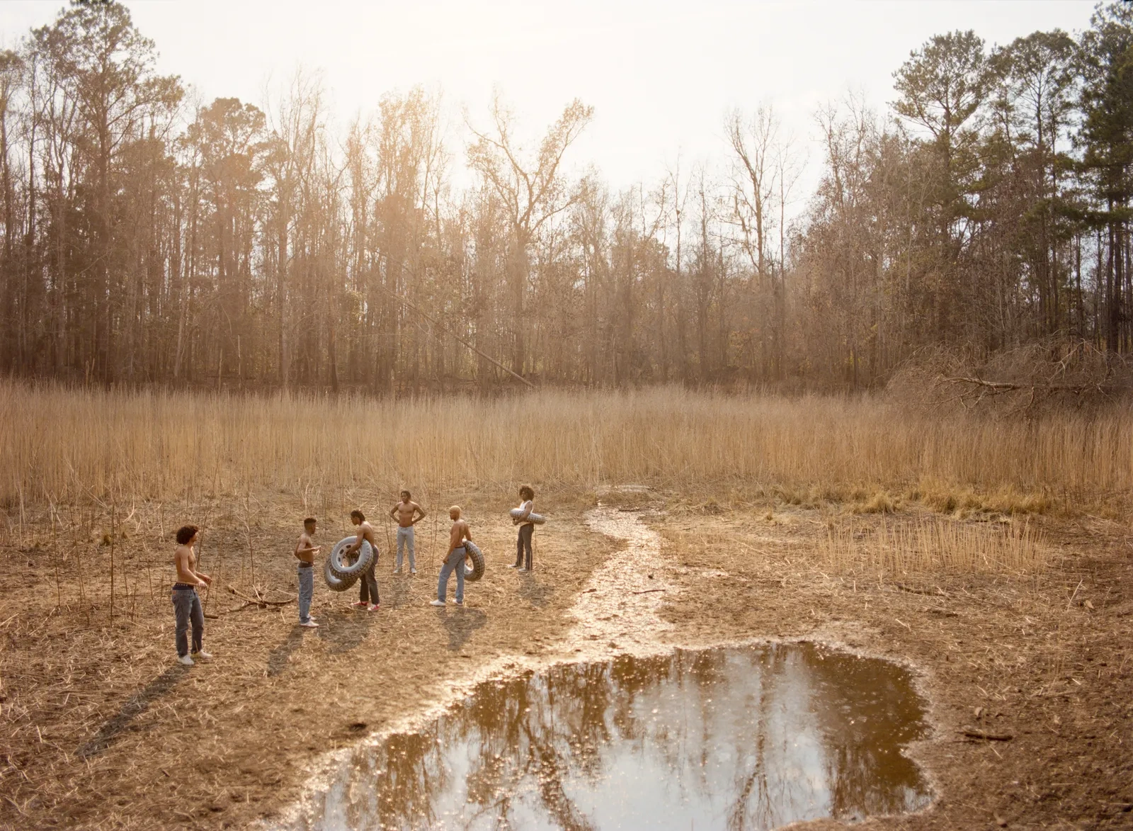 Florida Boys by Josh Aronson — A group of shirtless adolescents occupy a cleared, sandy field scattered with dried vegetation and shallow standing water.