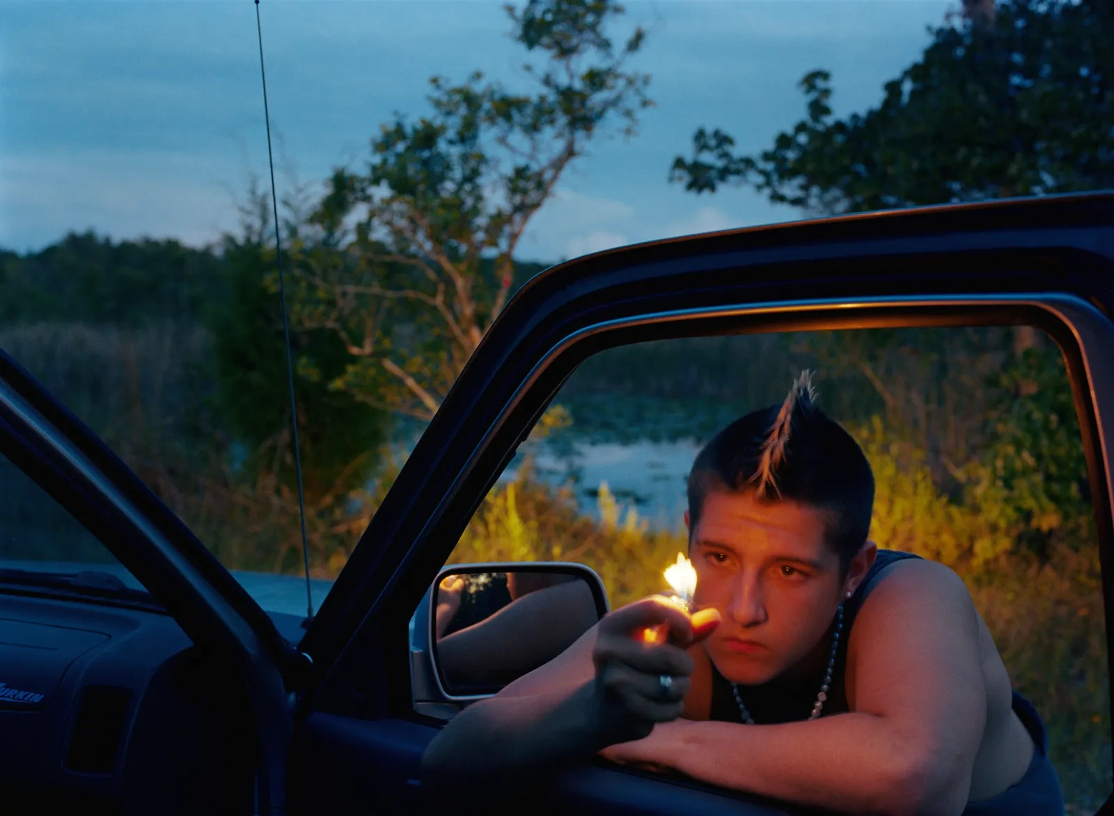 Florida Boys by Josh Aronson — A young man leans from the open window of a blue truck at dusk, his silhouette framed against a forested landscape and distant water.