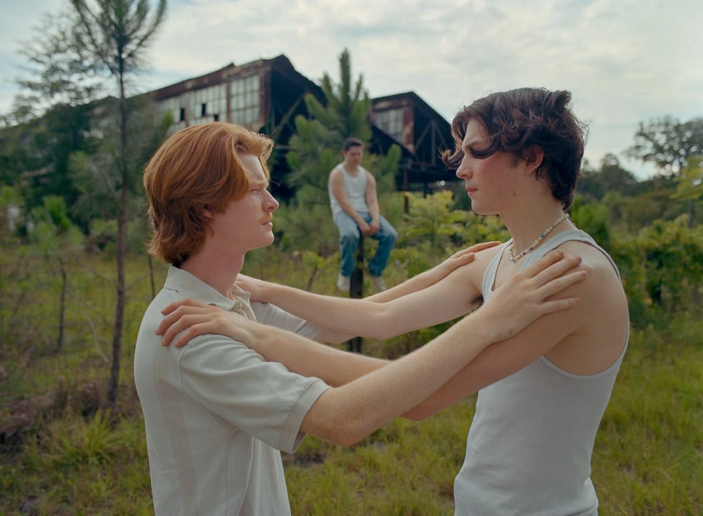 Florida Boys by Josh Aronson — Two young men in white tank tops engage in close physical contact within a scrubland landscape dotted with young pine saplings and tall dried grass.