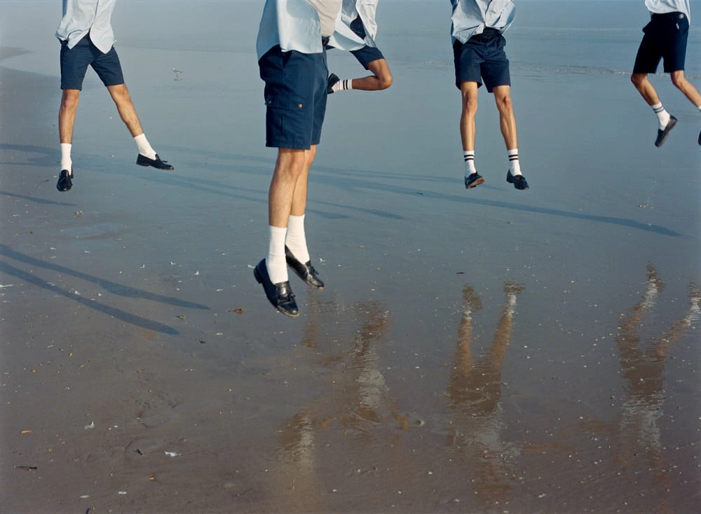Florida Boys by Josh Aronson — Five young men in white shirts and navy shorts run across wet sand and shallow water, their bodies captured mid-stride in varying states of suspension.