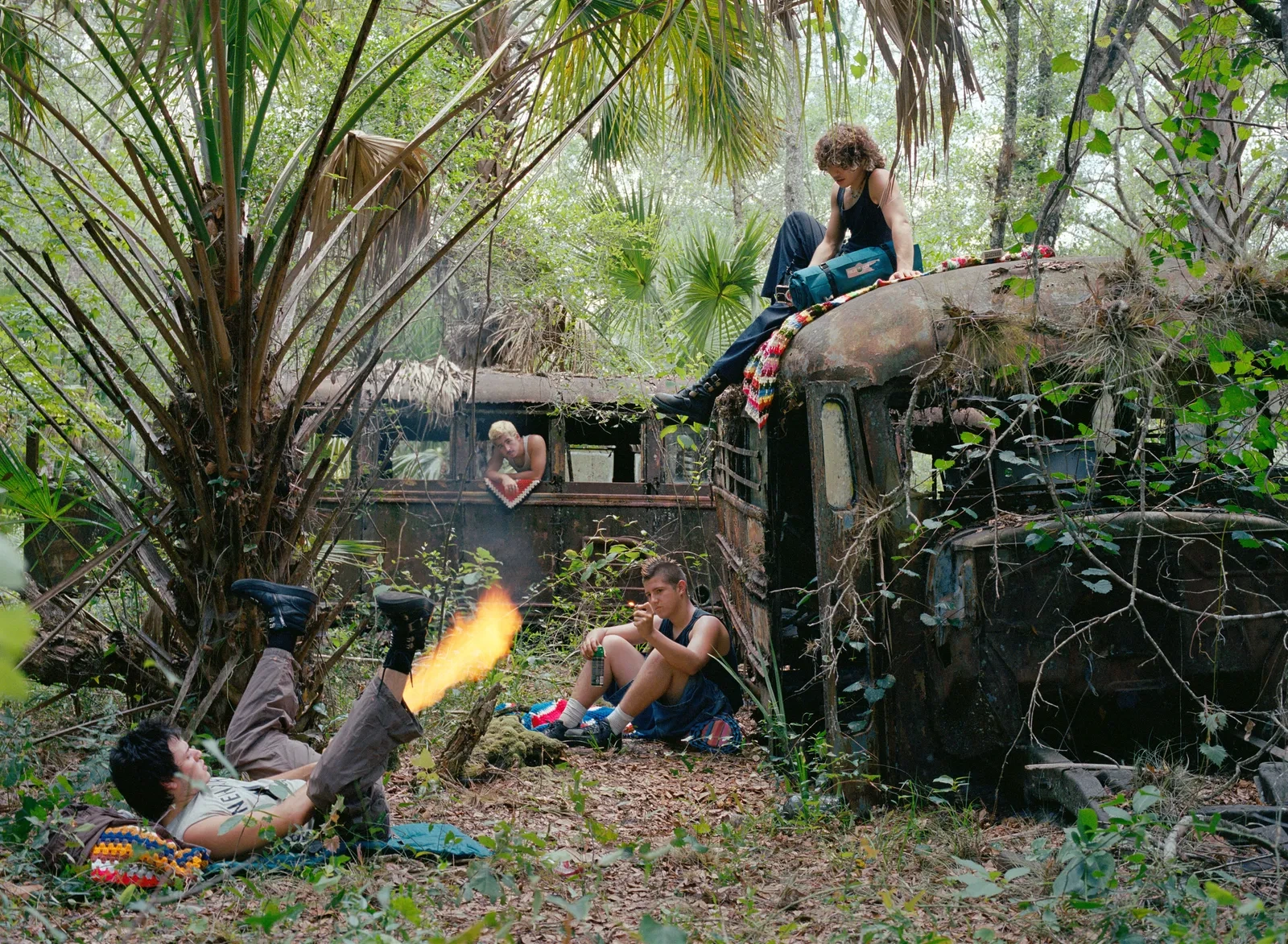 Florida Boys by Josh Aronson — In this scene from Florida Boys, three figures occupy a derelict truck consumed by subtropical overgrowth, its metal chassis corroded and reclaimed by vines...
