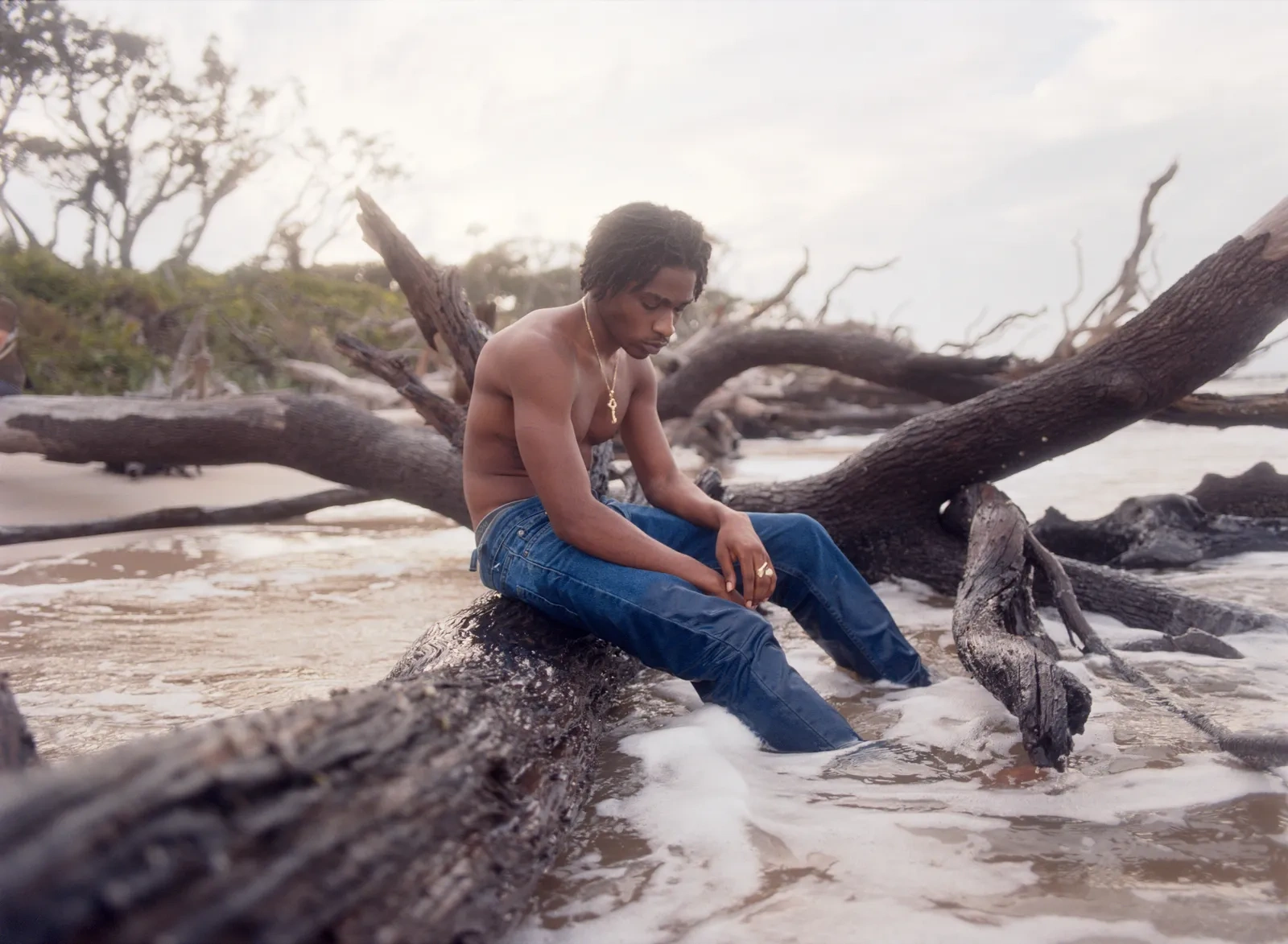Florida Boys by Josh Aronson — A young man sits on weathered driftwood at the shoreline, his posture introspective as he gazes downward.
