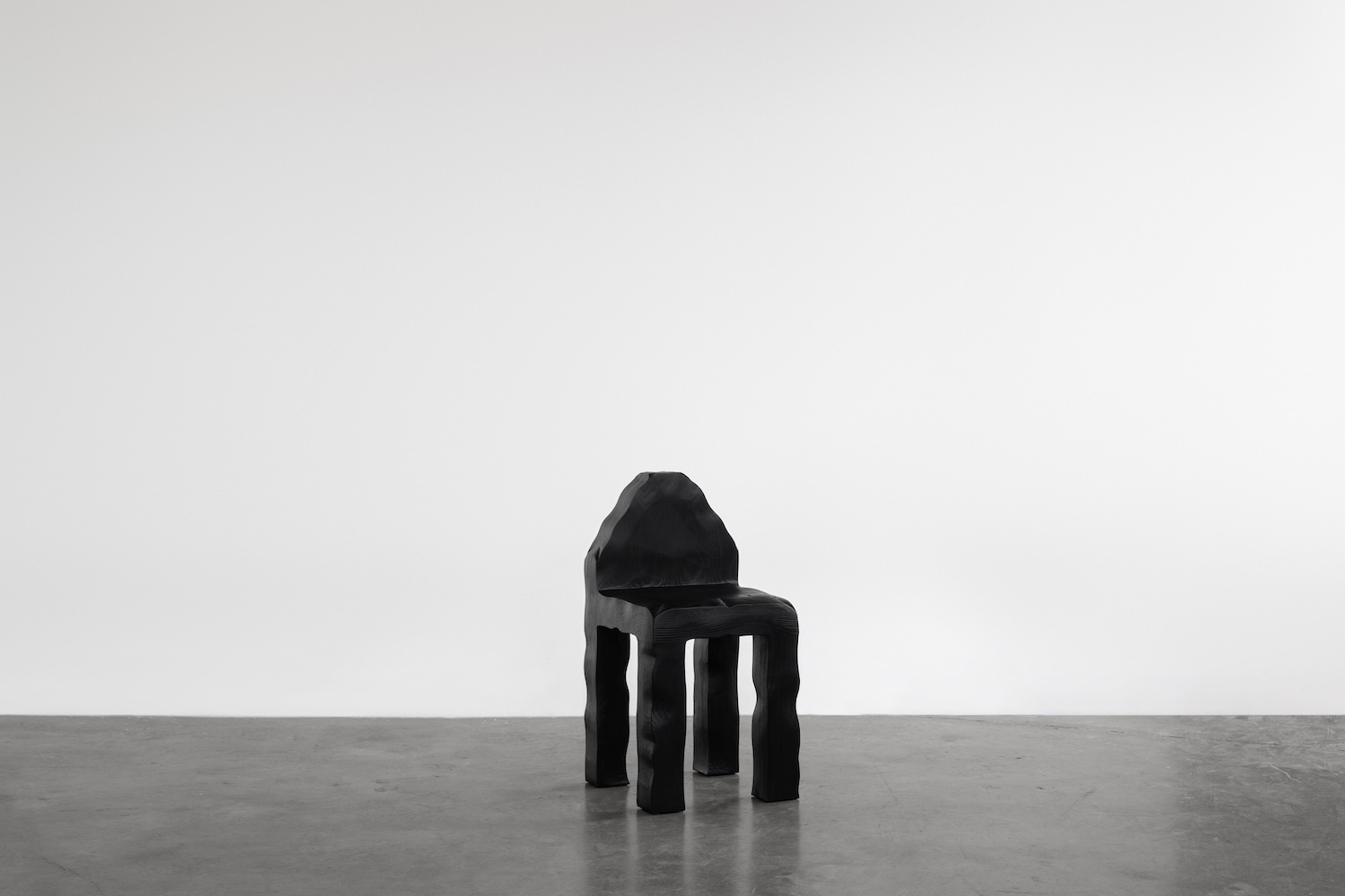 KAMEH 0.5 Collection by KAMEH — A charred timber chair from the KAMEH 0.5 collection sits centered on a concrete floor, its Shou Sugi Ban surface revealing pronounced wood grain striations...