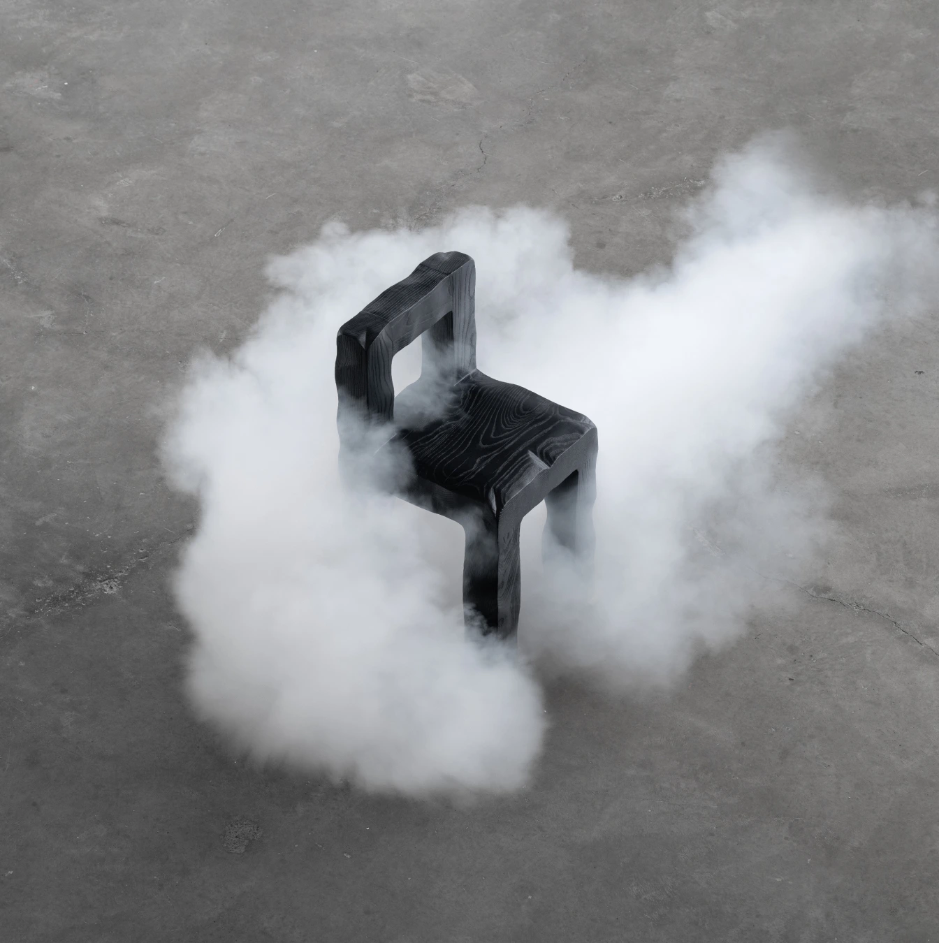 KAMEH 0.5 Collection by KAMEH — A charred timber chair from the KAMEH 0.5 collection emerges from dense white smoke on a concrete floor, captured from directly overhead.