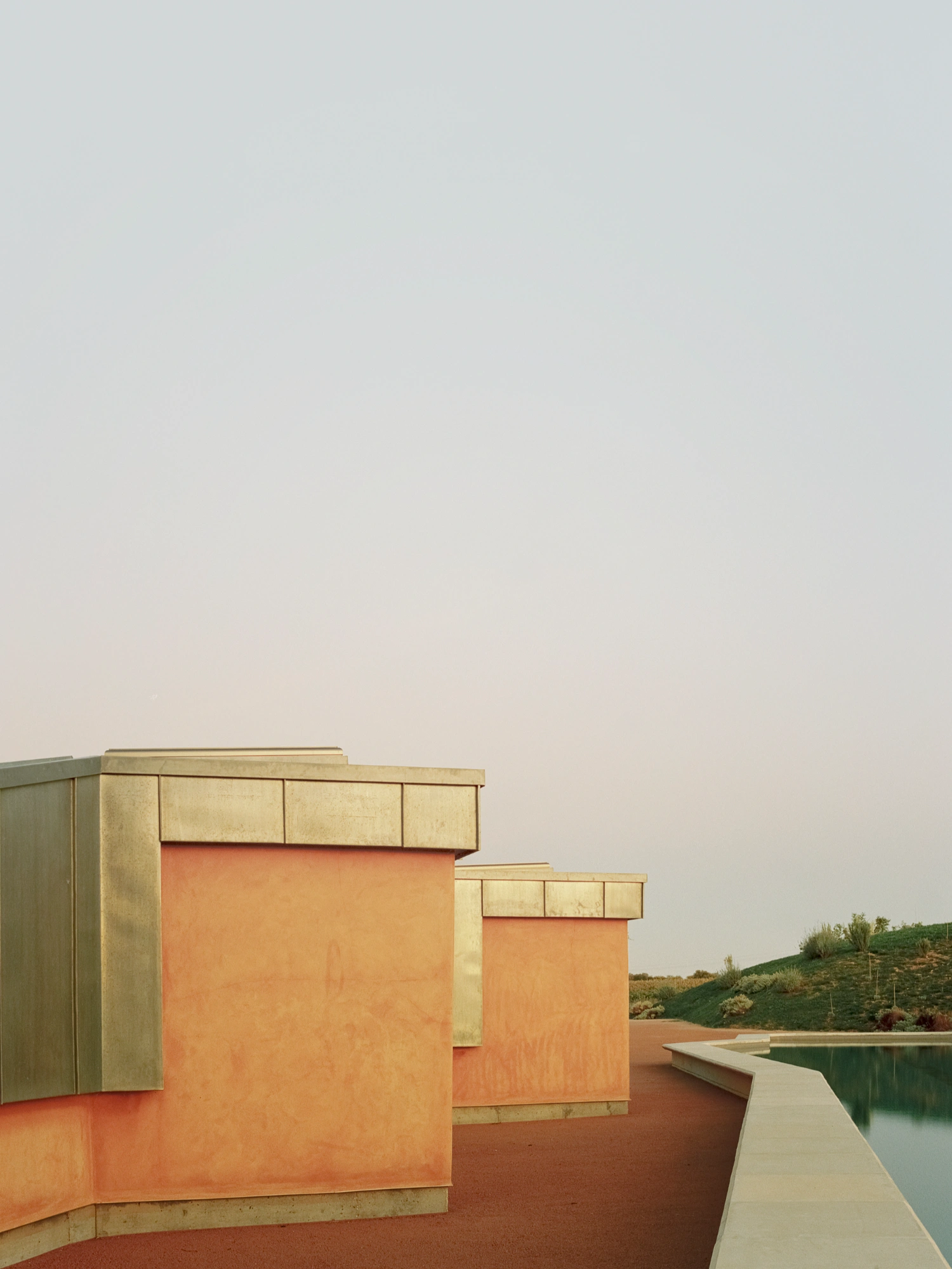 Herdade da Cardeira Winery by EMBAIXADA by EMBAIXADA arquitectura — Stepped volumetric forms in terracotta render rise against a pale sky, their horizontal bands of patinated metal cladding marking material transitions.