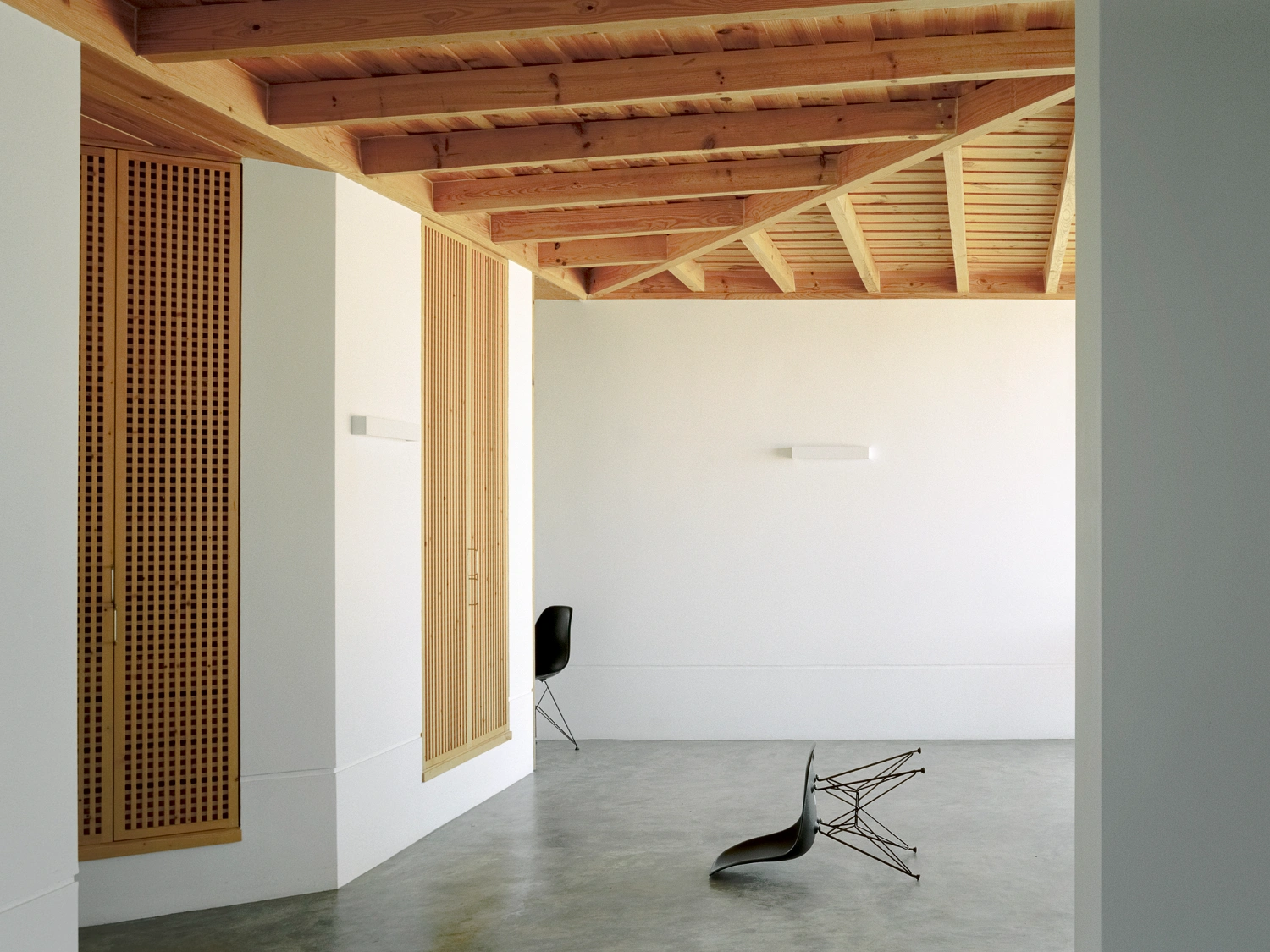 Herdade da Cardeira Winery by EMBAIXADA by EMBAIXADA arquitectura — A spacious interior gallery beneath exposed timber beams showcases two sculptural chairs with skeletal metal frames and curved black seats positioned on...