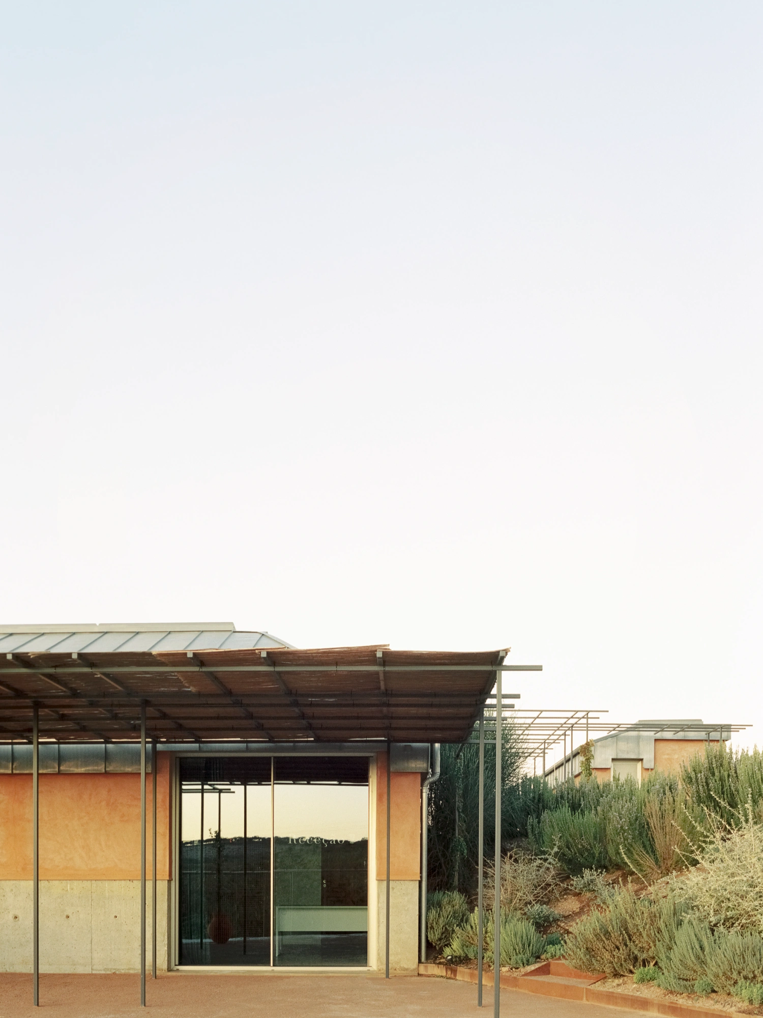 Herdade da Cardeira Winery by EMBAIXADA by EMBAIXADA arquitectura — A low-angle frontal view of the winery's primary pavilion reveals the material stratification governing the facade: terracotta-colored render occupies the...