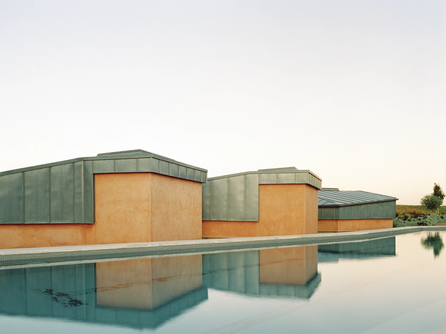 Herdade da Cardeira Winery by EMBAIXADA by EMBAIXADA arquitectura — The winery's paired pavilions present a studied material dialogue: oxidized copper or patinated metal cladding forms the upper horizontal bands, while...