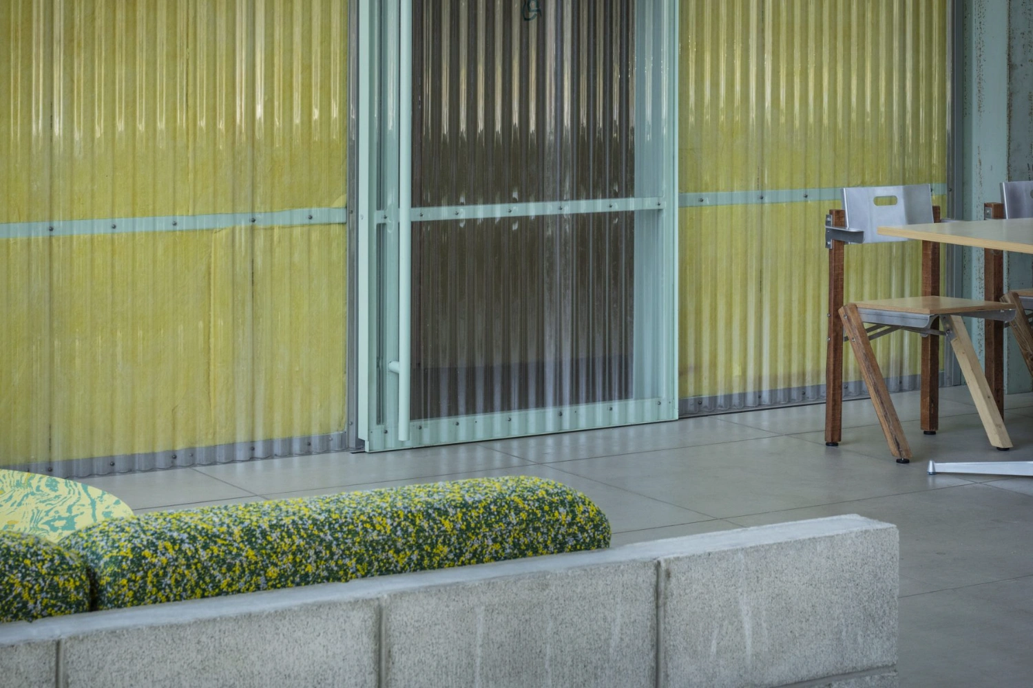 Teshima Factory Cafe by Schemata Architects — The cafe interior frames a composition of material contrasts: a raw concrete bench with yellow-and-grey textured upholstery anchors the foreground, while...