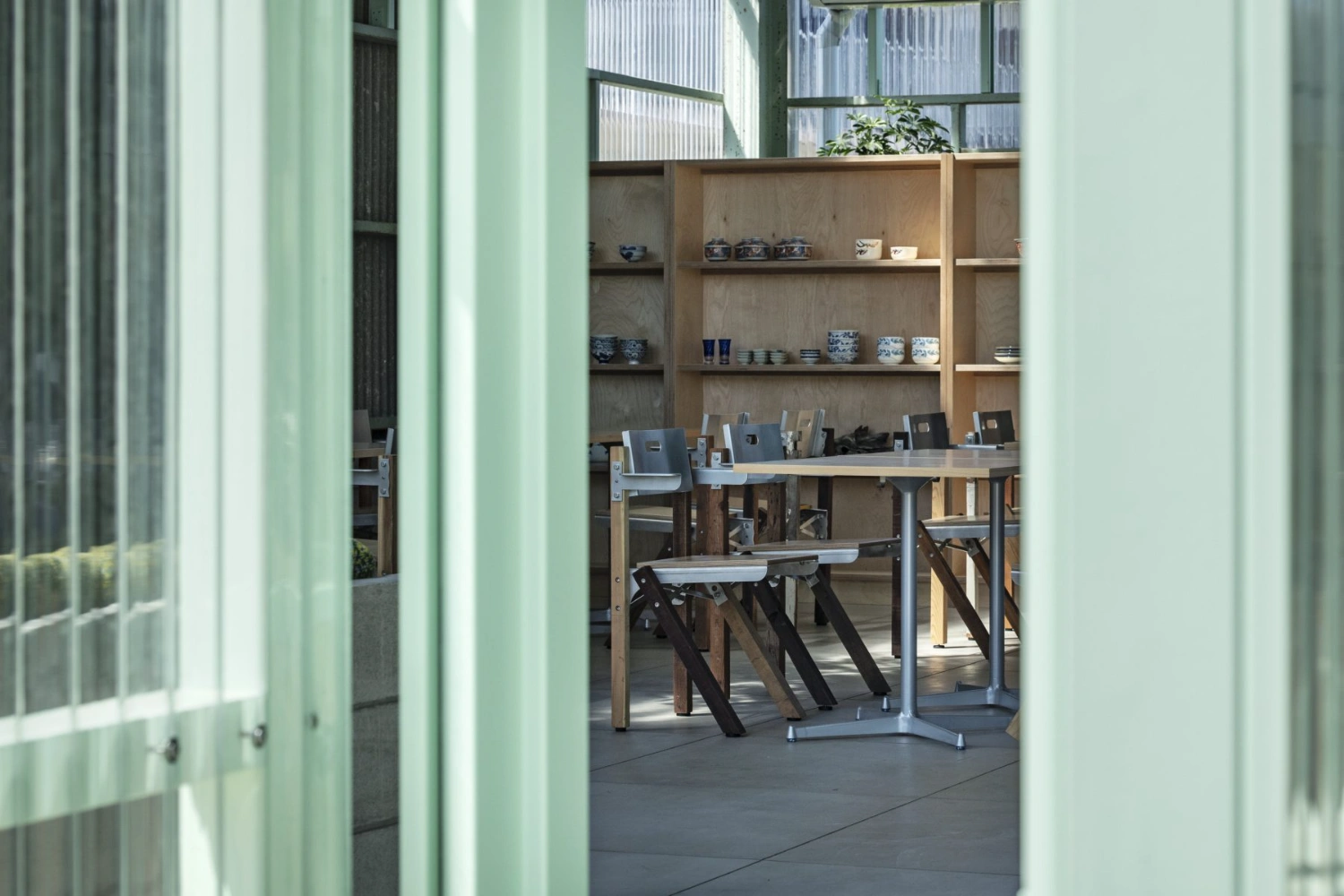 Teshima Factory Cafe by Schemata Architects — View through sage-green translucent corrugated panels into the café interior, where timber shelving systems display ceramic vessels and tableware against a...