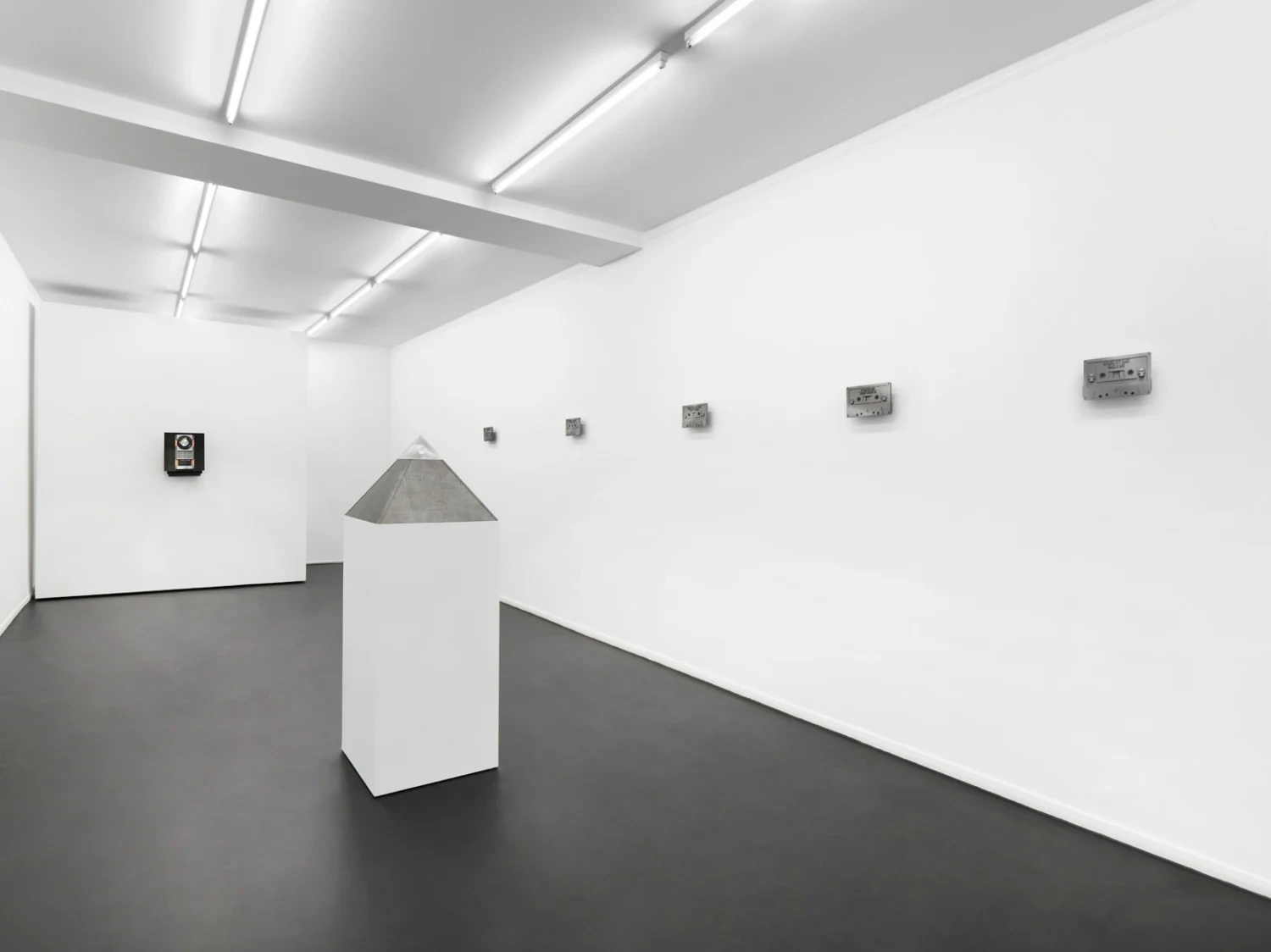 “Heaven’s worth, Hell on earth” by Christelle Oyiri at Galerie Bucholz — A monolithic gray concrete pyramid mounted on a white plinth bisects the gallery floor, its textured surface contrasting sharply against pristine walls.