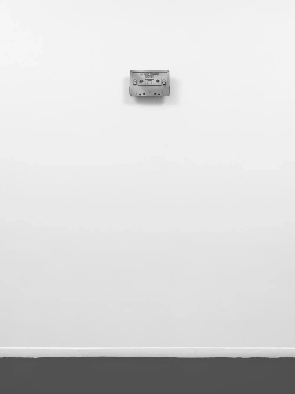 “Heaven’s worth, Hell on earth” by Christelle Oyiri at Galerie Bucholz — A wall-mounted metallic cassette-like device sits centered on white gallery wall, its rectangular form featuring dual reels and control mechanisms visible...