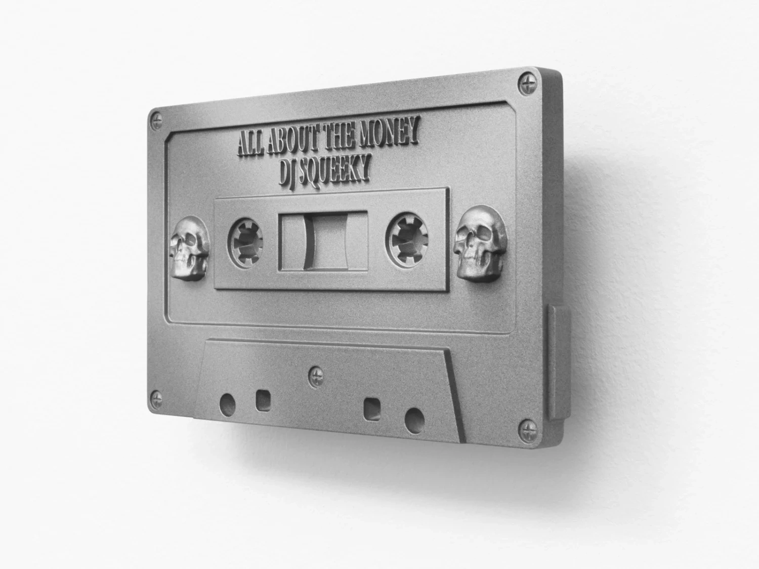 “Heaven’s worth, Hell on earth” by Christelle Oyiri at Galerie Bucholz — A cast aluminum cassette tape dominates the composition, rendered in monochromatic silver-gray with embossed typography reading 'ALL ABOUT THE MONEY DJ...