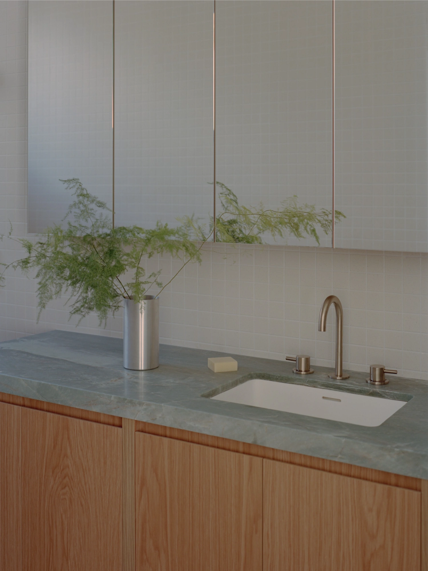 Mount Martha House by Victoria Merrett Architects — A bathroom vanity in pale green-grey stone anchors the composition, its matte surface supporting a cylindrical stainless steel vessel containing feathery...