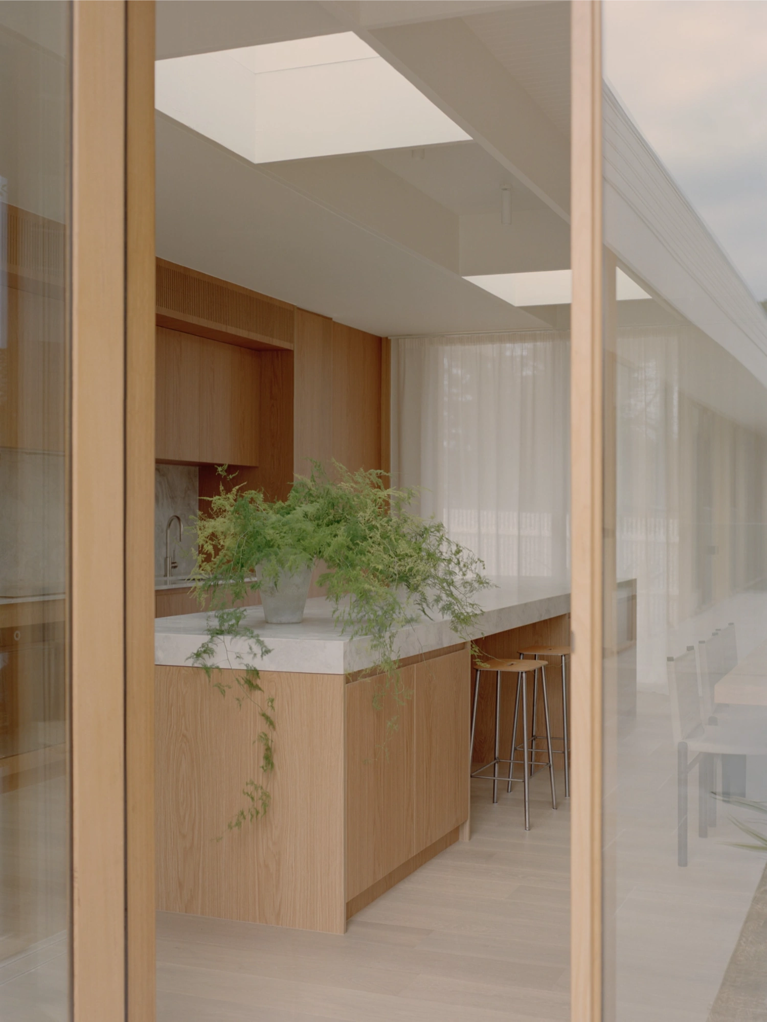 Mount Martha House by Victoria Merrett Architects — A kitchen viewed through a timber-framed glass threshold, its open-plan layout defined by a timber island with white countertop and integrated planter.