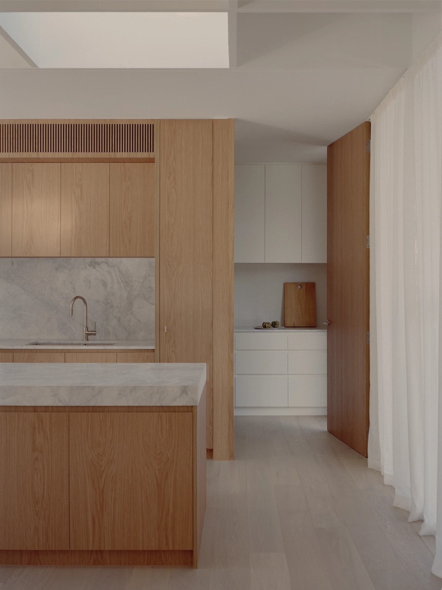 Mount Martha House by Victoria Merrett Architects — The kitchen and circulation space reveals the project's material palette in sequence: warm oak cabinetry with vertical slat detailing transitions to white...