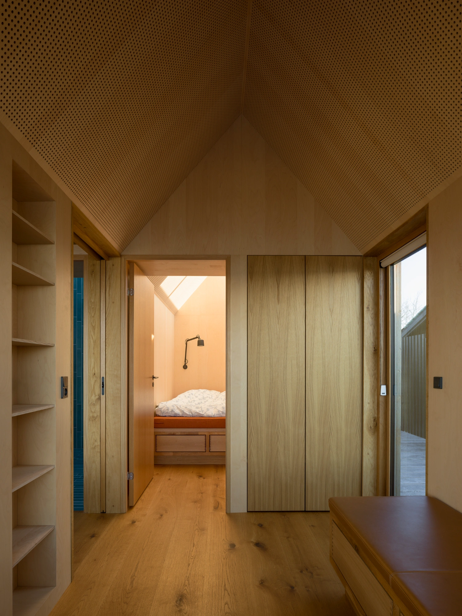 Strandvejen 35 Summer House by Jesper Kusk Arkitekter — Interior corridor of one gabled volume, lined with built-in oak shelving on the left and paired sliding doors clad in honey-toned wood grain on the right.