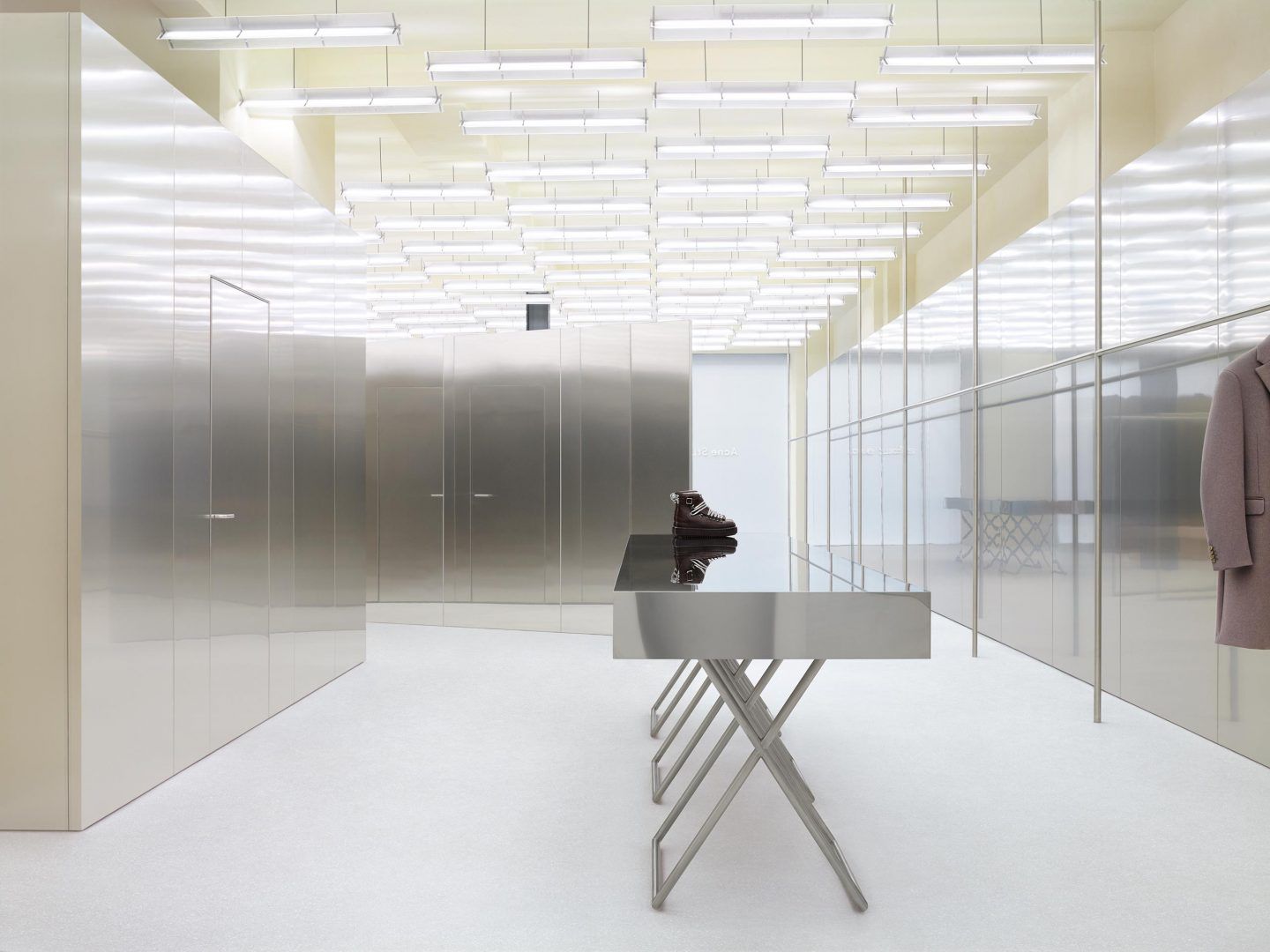Acne Studios by Bubab Architects — A centrally positioned stainless steel X-frame table with rectangular top anchors the narrow retail corridor.