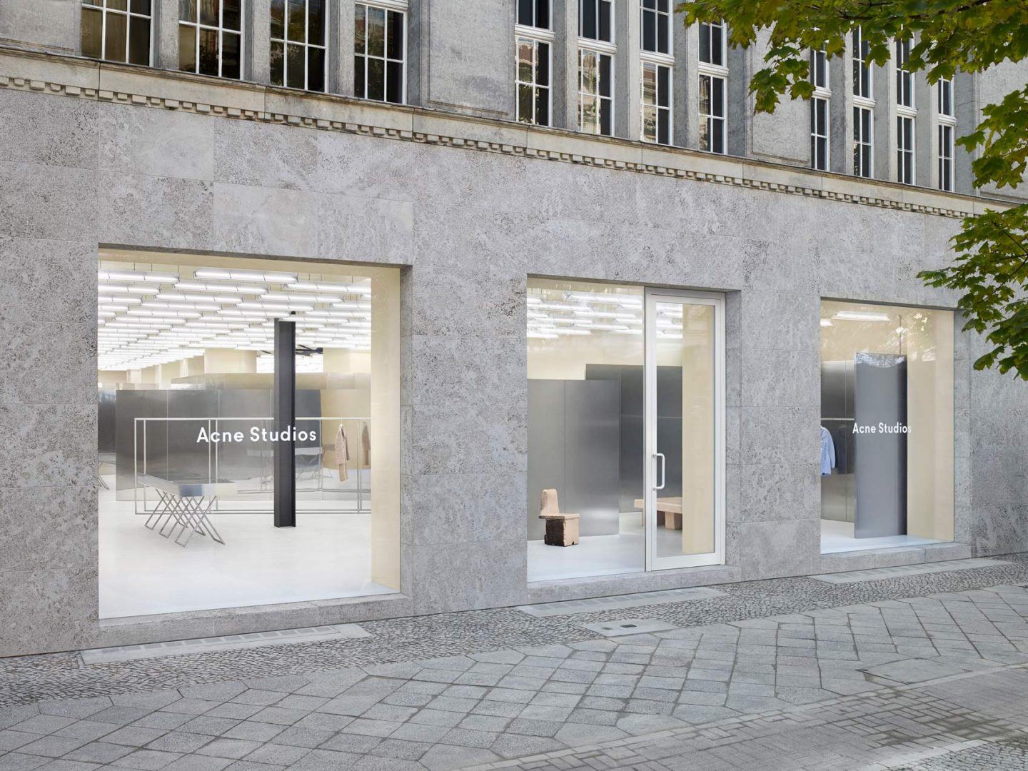 Acne Studios by Bubab Architects — Street-level view of Acne Studios Berlin storefront reveals a tripartite facade of floor-to-ceiling glazing separated by pale concrete piers.