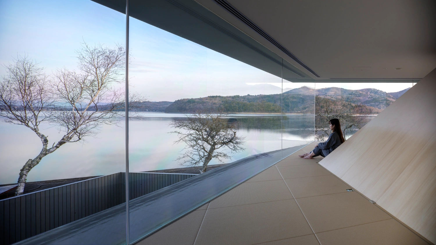 CYCL by YŪ Momoeda Architects — A solitary figure sits at the edge of a floor-to-ceiling frameless glass plane, overlooking Yamanakako's still water and forested shoreline.