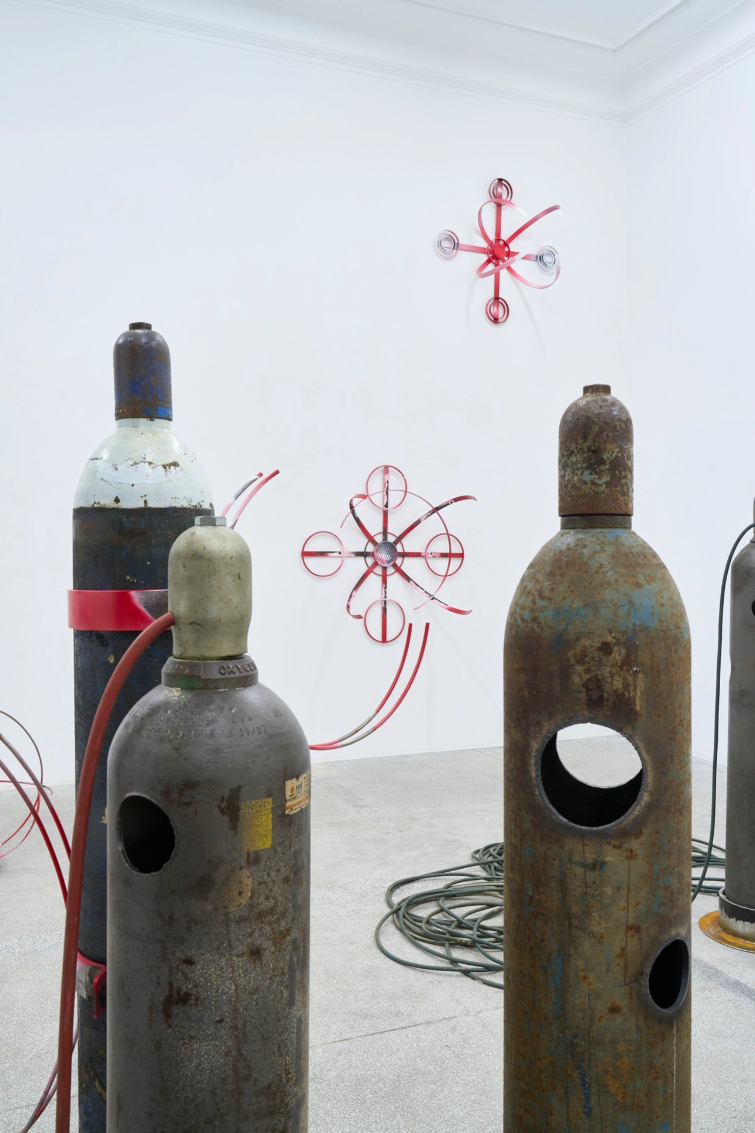 Swarm / Rój by Zuza Golinska at Piktogram — Industrial gas cylinders with perforated circular apertures occupy the gallery floor, their oxidized metal surfaces—rusted orange,...