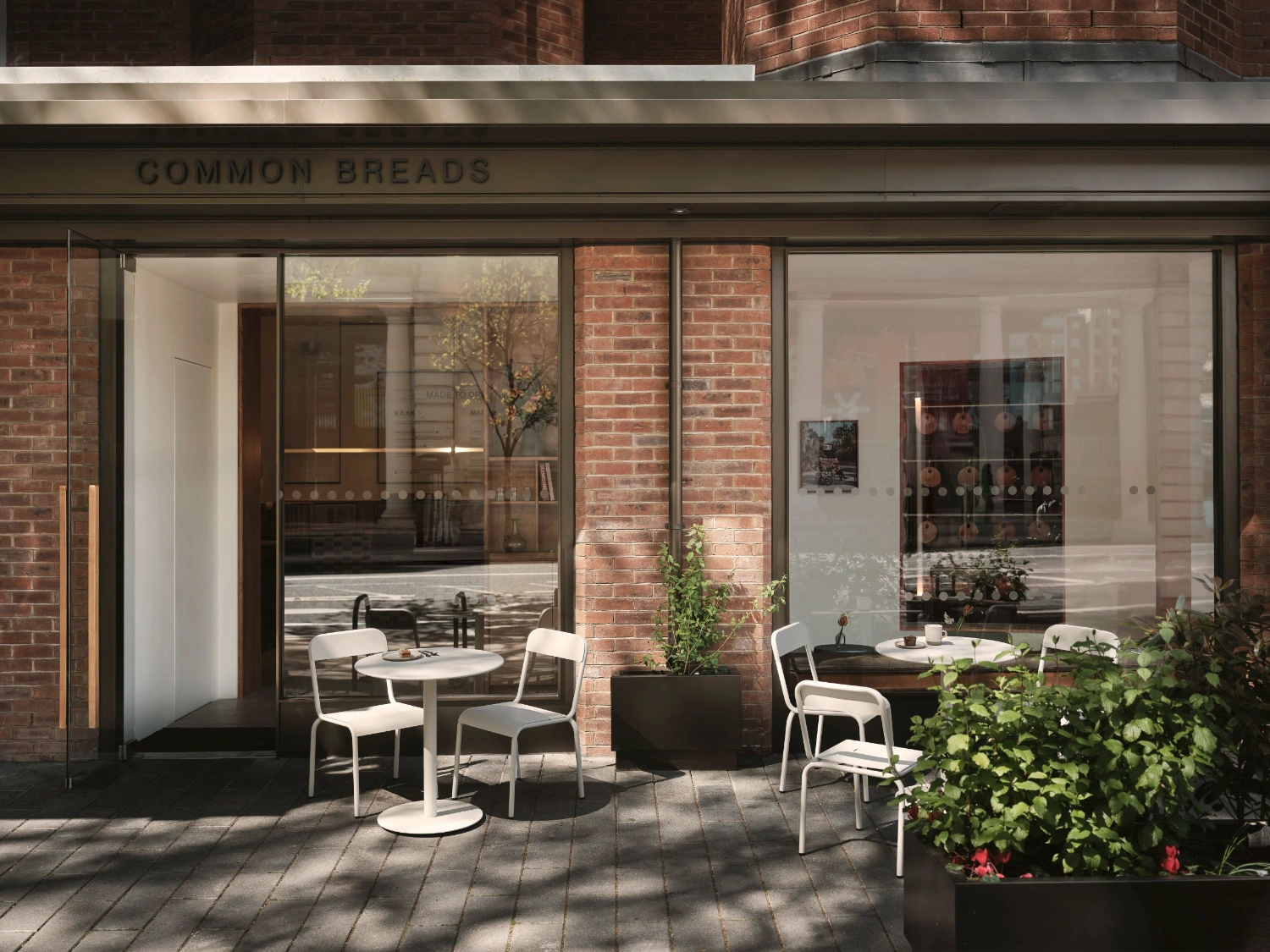 Common Breads bakery by MA Studio — Common Breads' street-facing courtyard showcases a minimalist dining arrangement beneath the storefront's dark-framed glazing.
