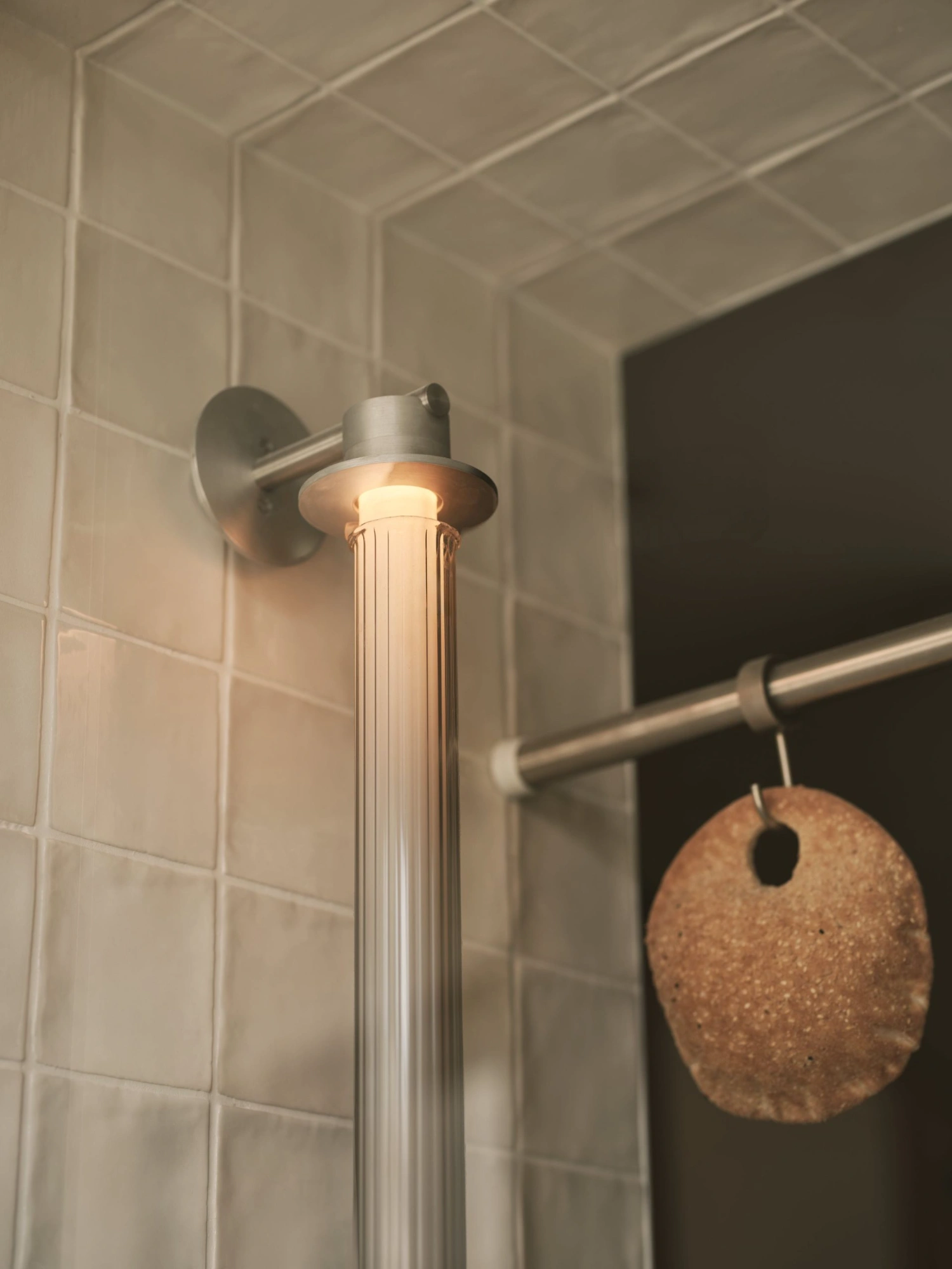Common Breads bakery by MA Studio — A close-up detail of the Common Breads bakery bathroom fixture showcases a brushed-metal pendant light with a cylindrical ribbed glass shade casting warm...