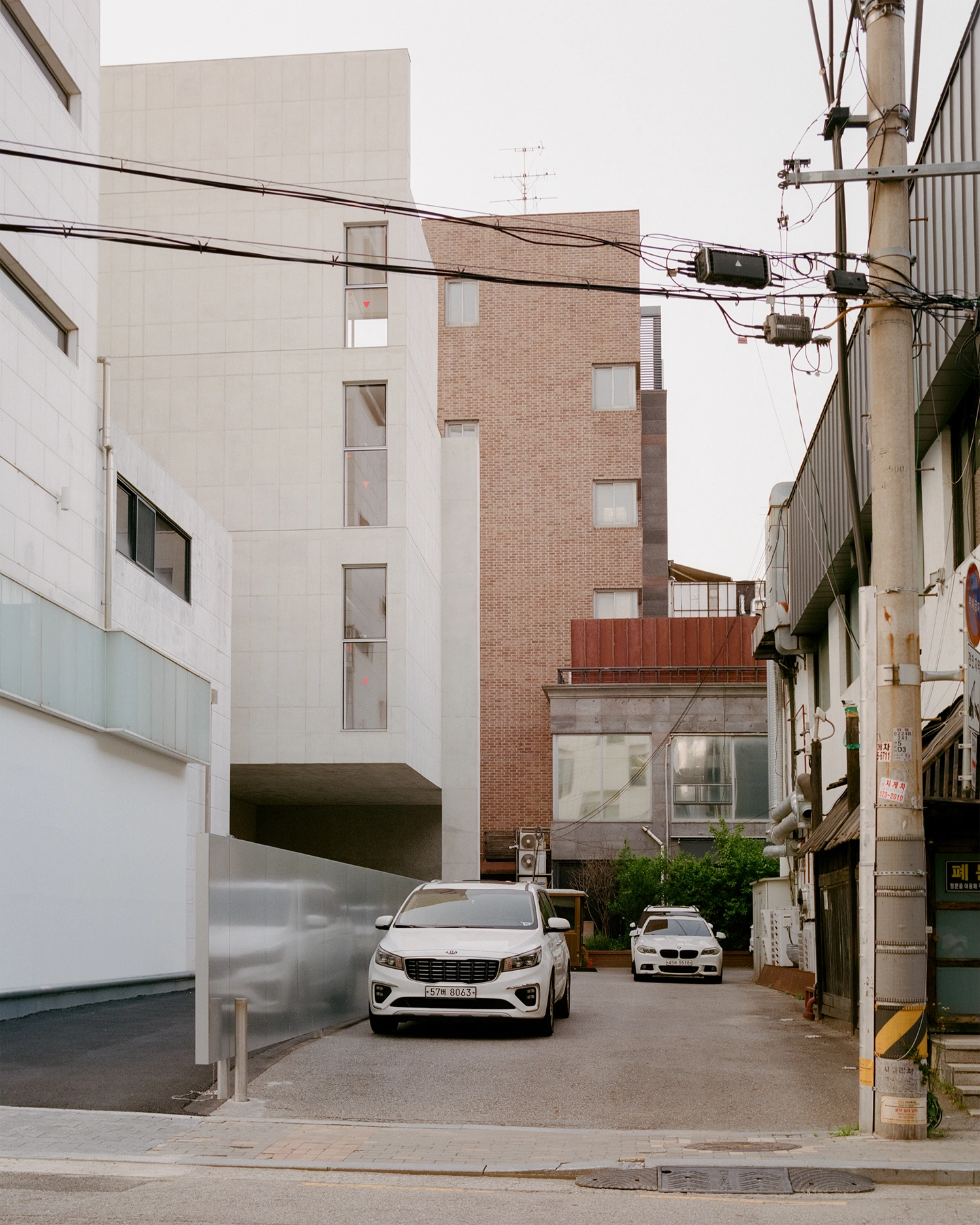 K-Auction Exhibition Hall Sinsa by min_workshop + 3ab — A narrow alleyway in Seoul frames the K-Auction Exhibition Hall's modernist facade, where a white vehicle is parked beneath a dramatic cantilevered concrete...